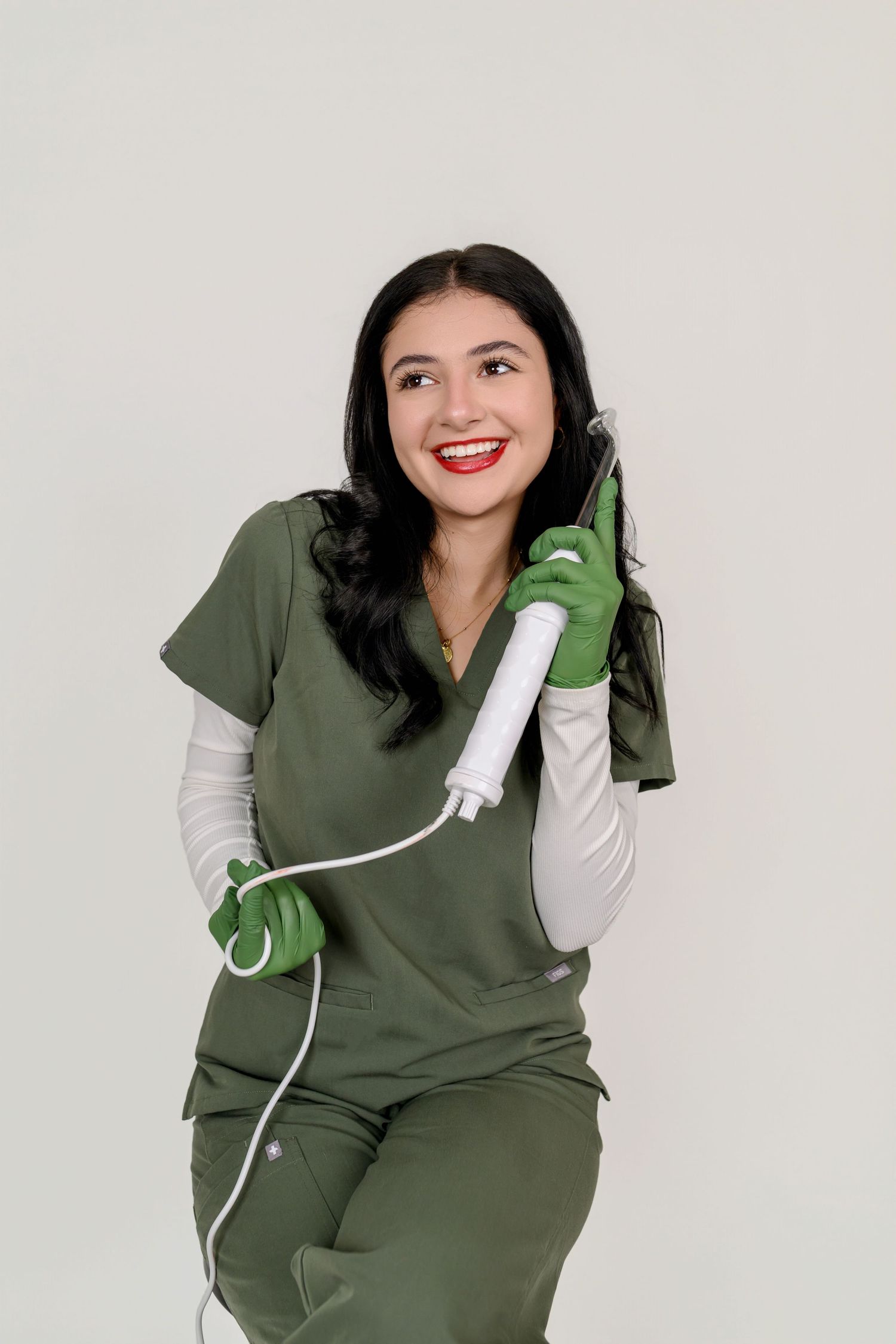 Smiling female medical professional in green scrubs holding a cosmetic device, wearing green gloves on white background.