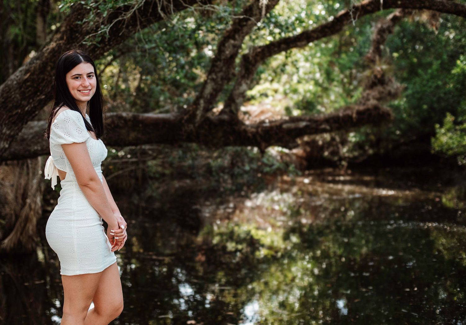 Someone in a white dress poses by a serene creek with overhanging trees reflecting in the calm water.