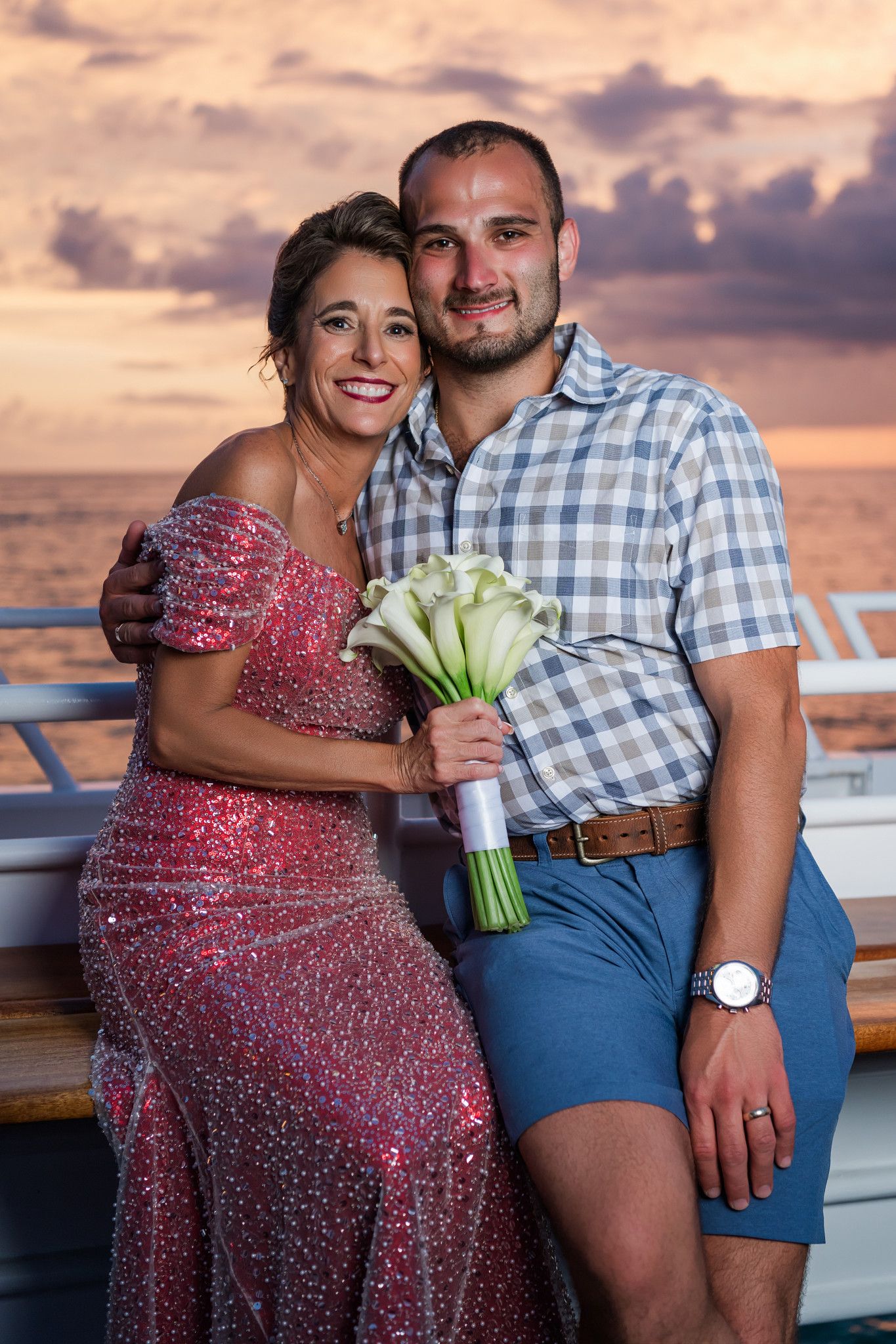 Couple posing on cruise ship deck at sunset, woman in pink sequin dress holding white calla lily bouquet, man in plaid shirt.