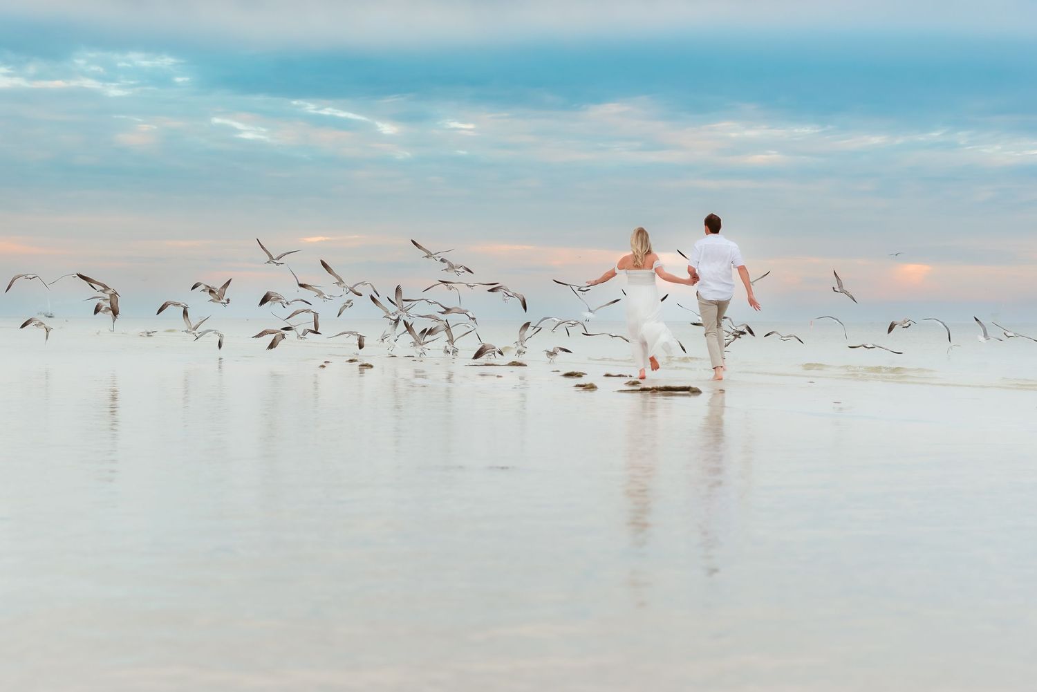 Couple in white walking on shallow water beach with seagulls flying around them at sunset.
