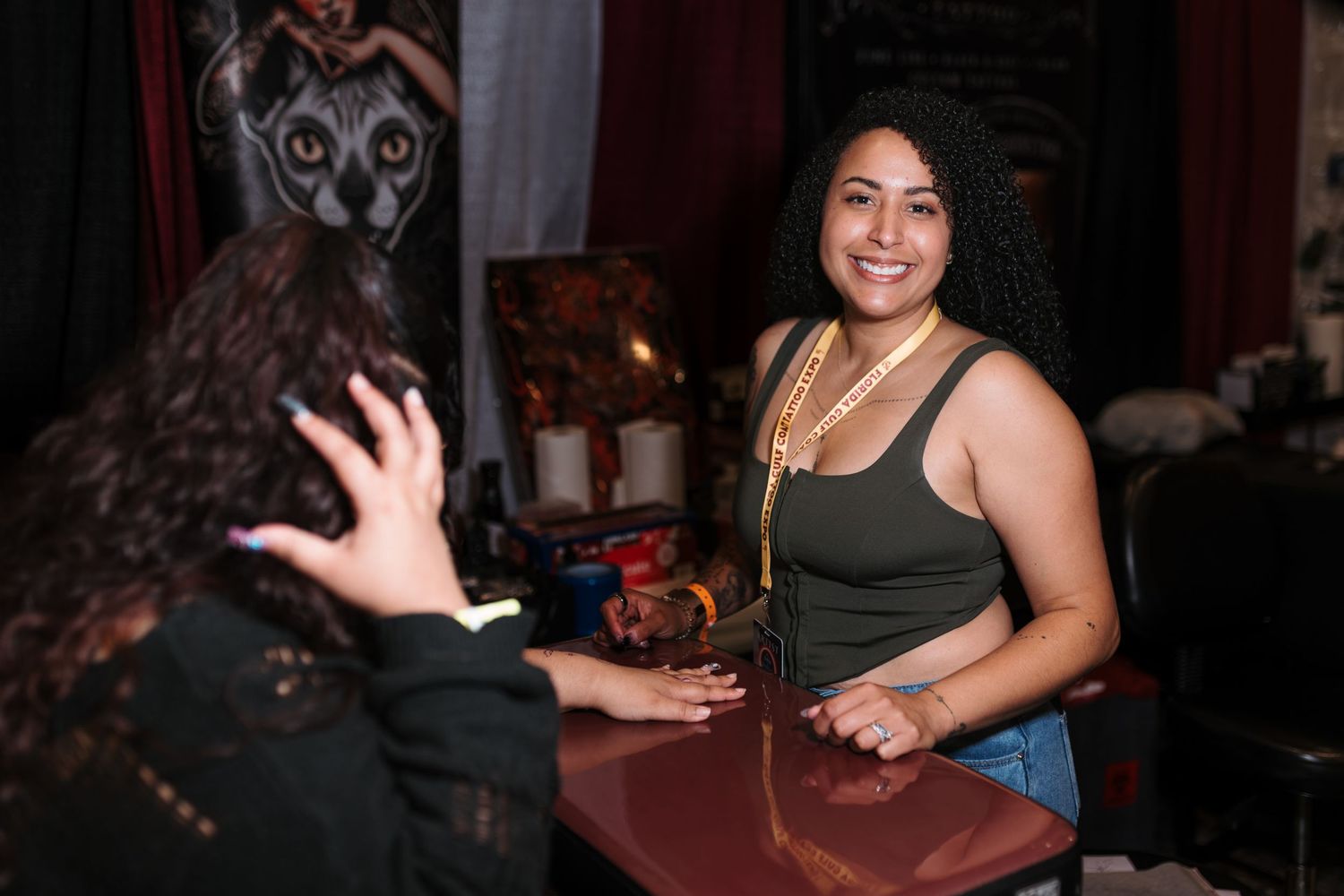 Two women smile and interact at a decorated vendor table at an indoor event or convention.