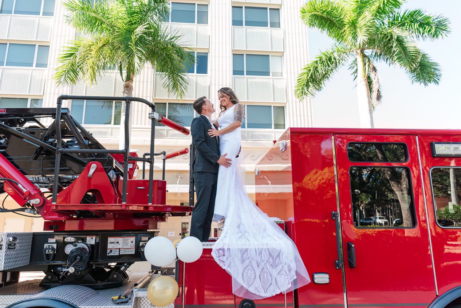 Newlyweds share a romantic embrace standing on top of a decorated red fire truck outside a building.
