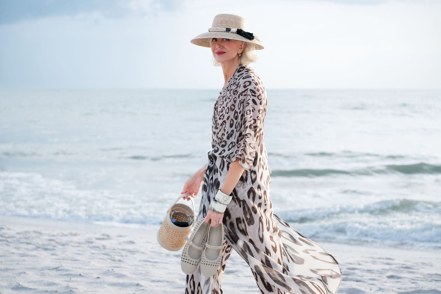 Woman in leopard print maxi dress and sun hat walking on a beach carrying a straw bag.