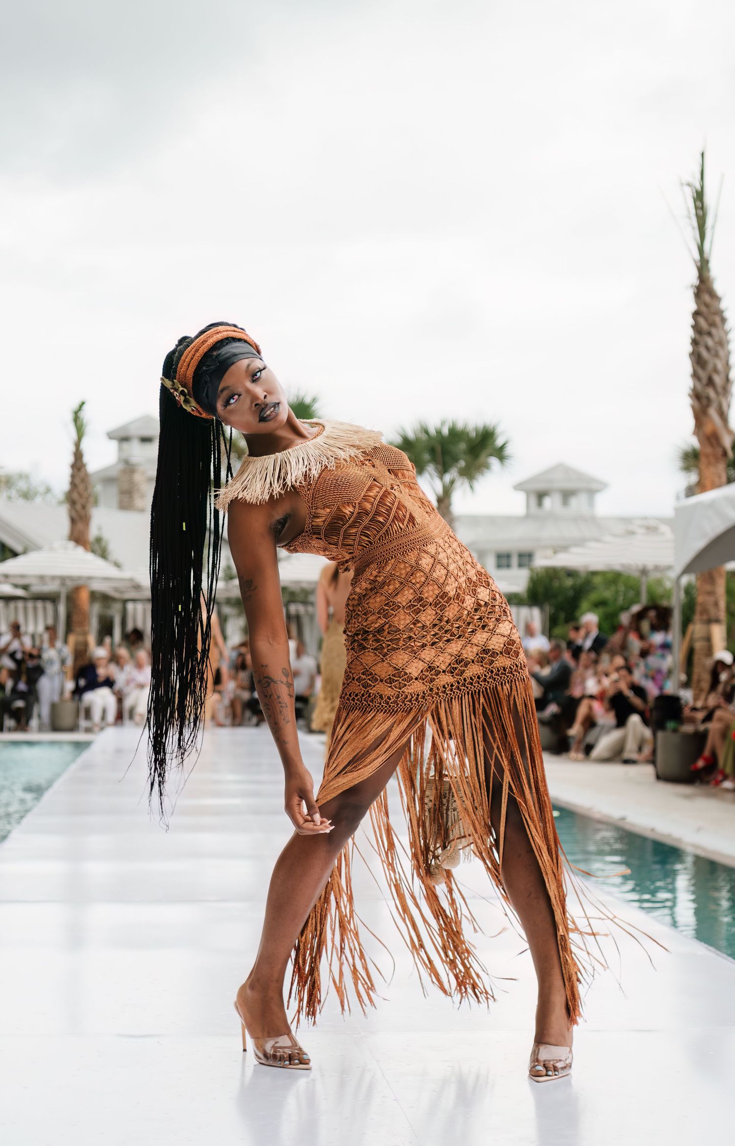 Model in brown fringe dress poses on runway near pool at outdoor fashion show.