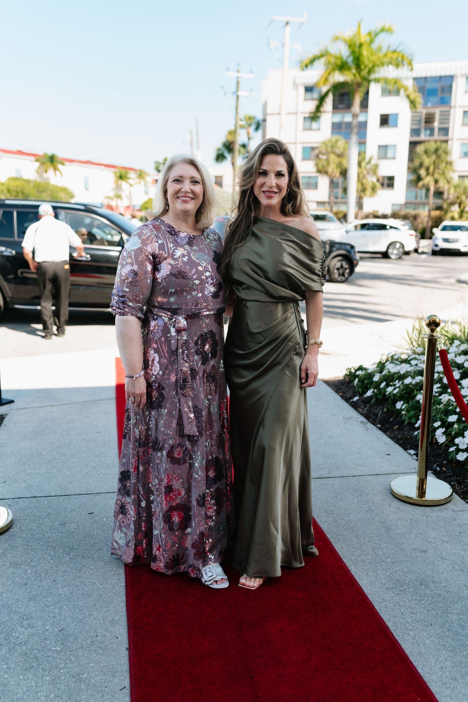 Two women in elegant evening gowns pose together on a red carpet at an outdoor event.