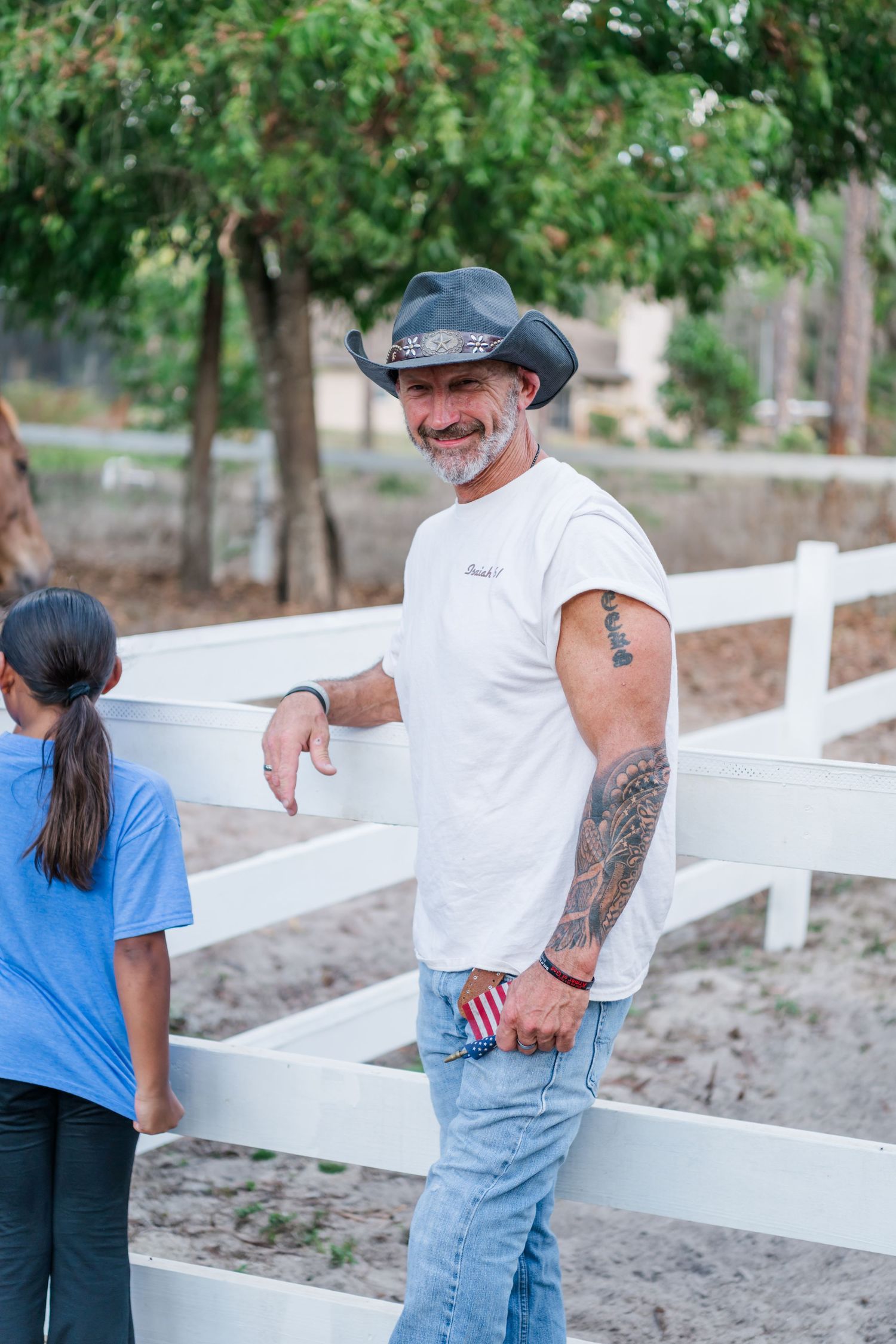 Two people painting a white wooden fence together in a backyard with trees in the background.