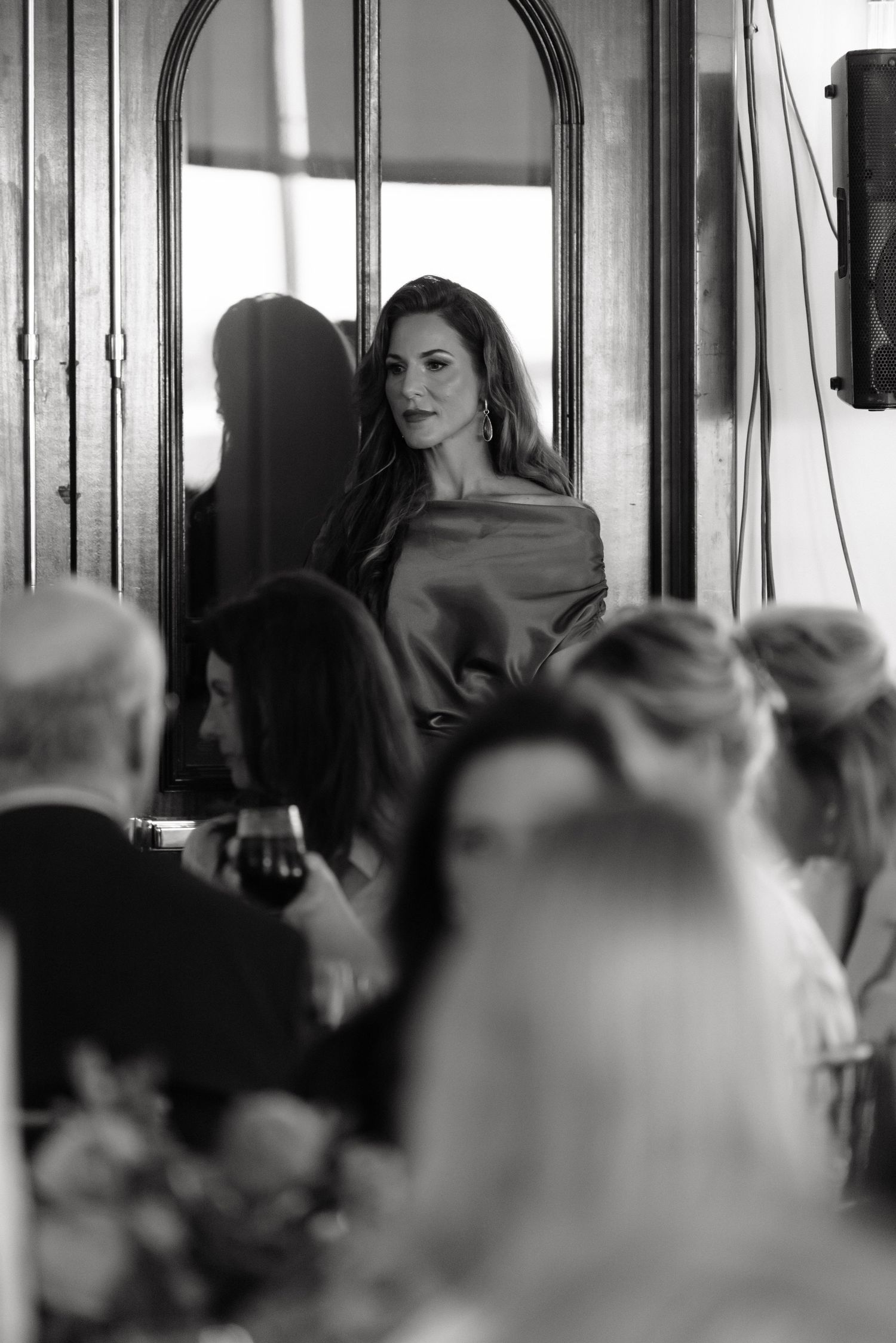 A black and white photograph shows an elegant figure in an off-shoulder dress standing in front of a window at an event.