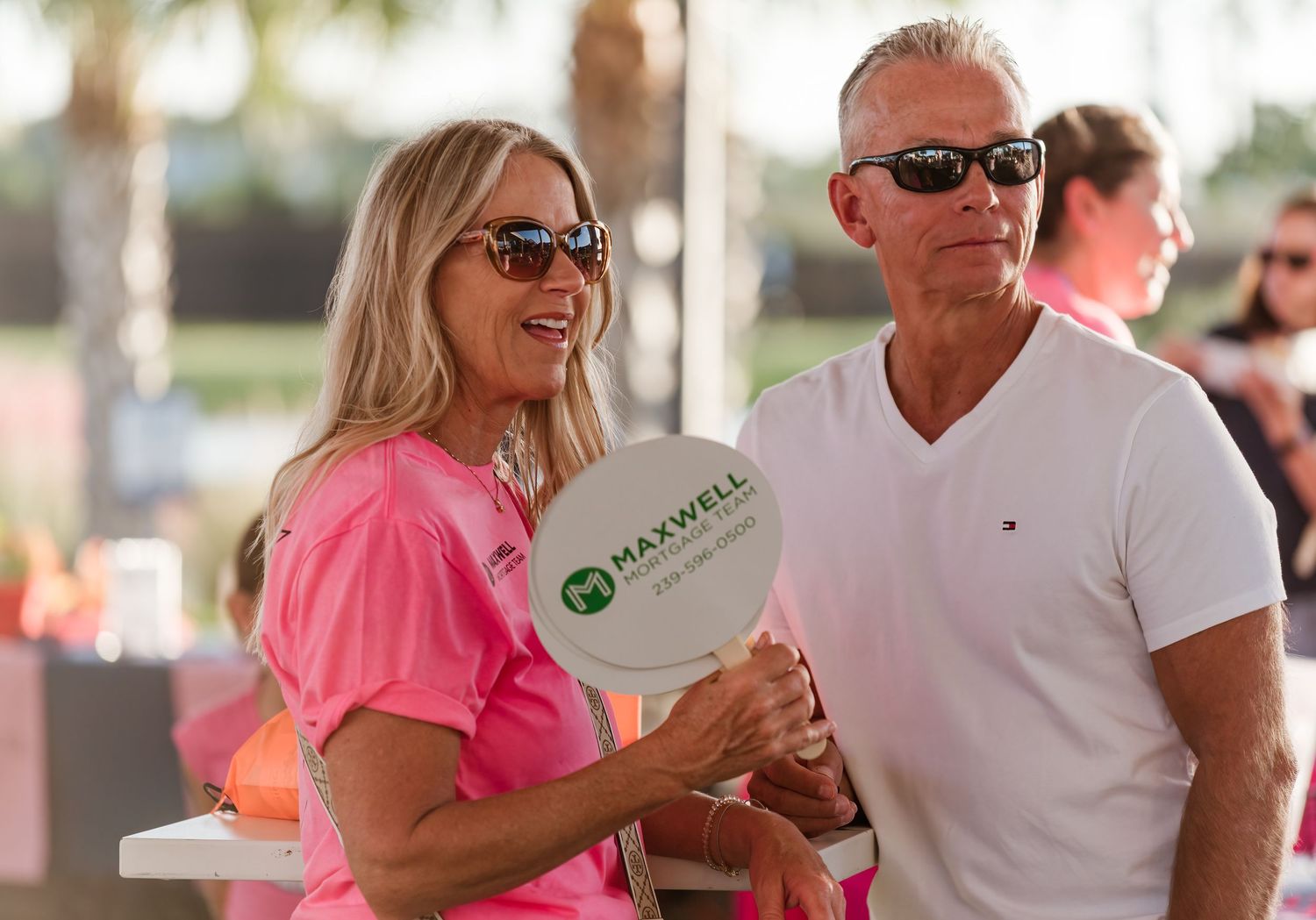 Two people in sunglasses at an outdoor event, with one wearing a pink shirt and holding a branded fan.