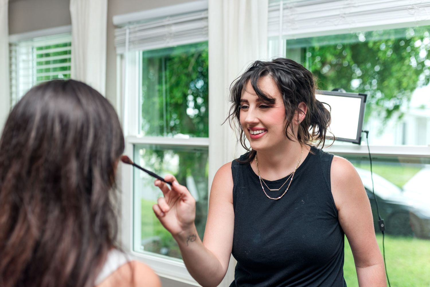 A makeup artist applies cosmetics using a brush while standing near a bright window with greenery visible outside.