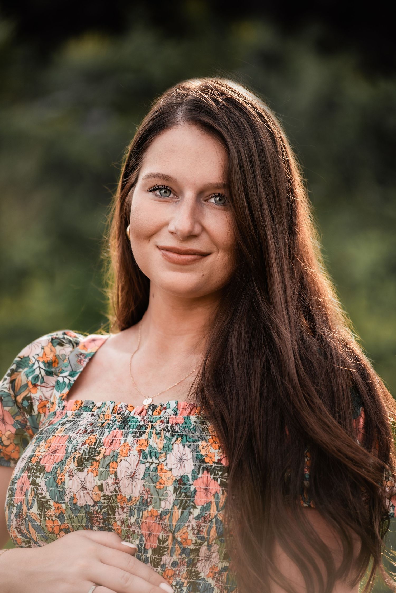 A person in a floral dress with long dark hair poses outdoors against a blurred natural background.