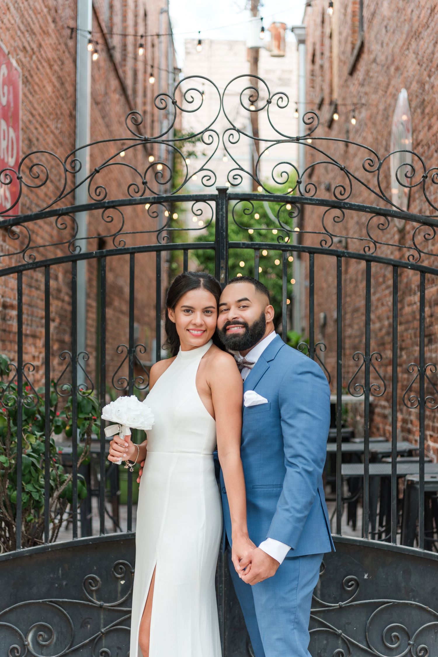 A couple poses in front of an ornate wrought iron gate between brick walls, with greenery visible through the gate.
