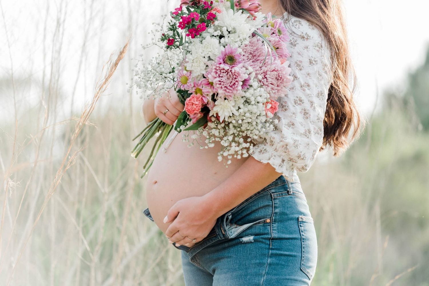 A pregnant woman in denim and lace holds a romantic bouquet of pink and white wildflowers in a meadow.