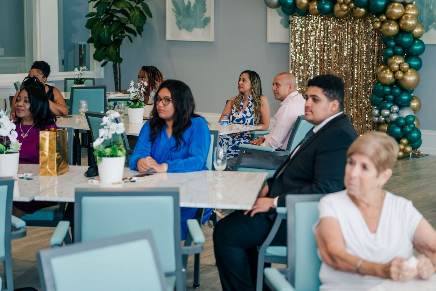 Guests seated at elegant tables during a celebration event with teal and gold balloon arch decor.
