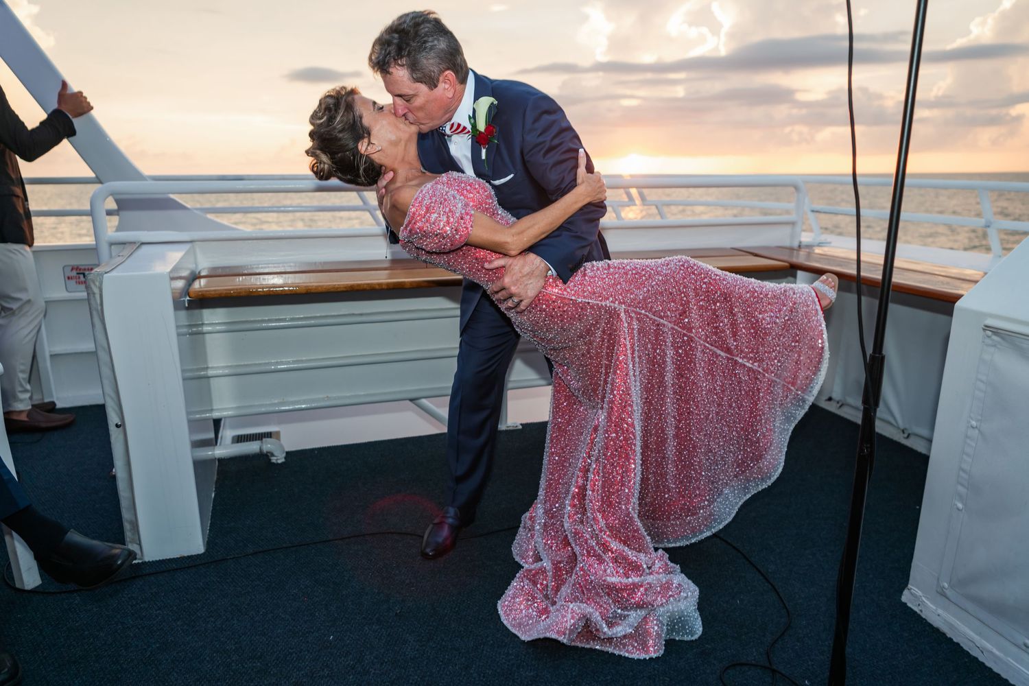 Couple sharing a romantic dip kiss on cruise ship deck at sunset, bride in pink gown and groom in navy suit.