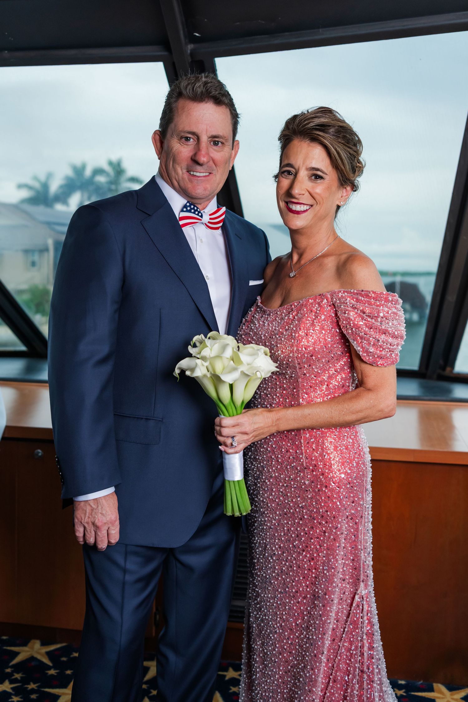 Couple posing in formal attire, man in navy suit with American flag bow tie, woman in pink sequin gown holding white calla lily bouquet.