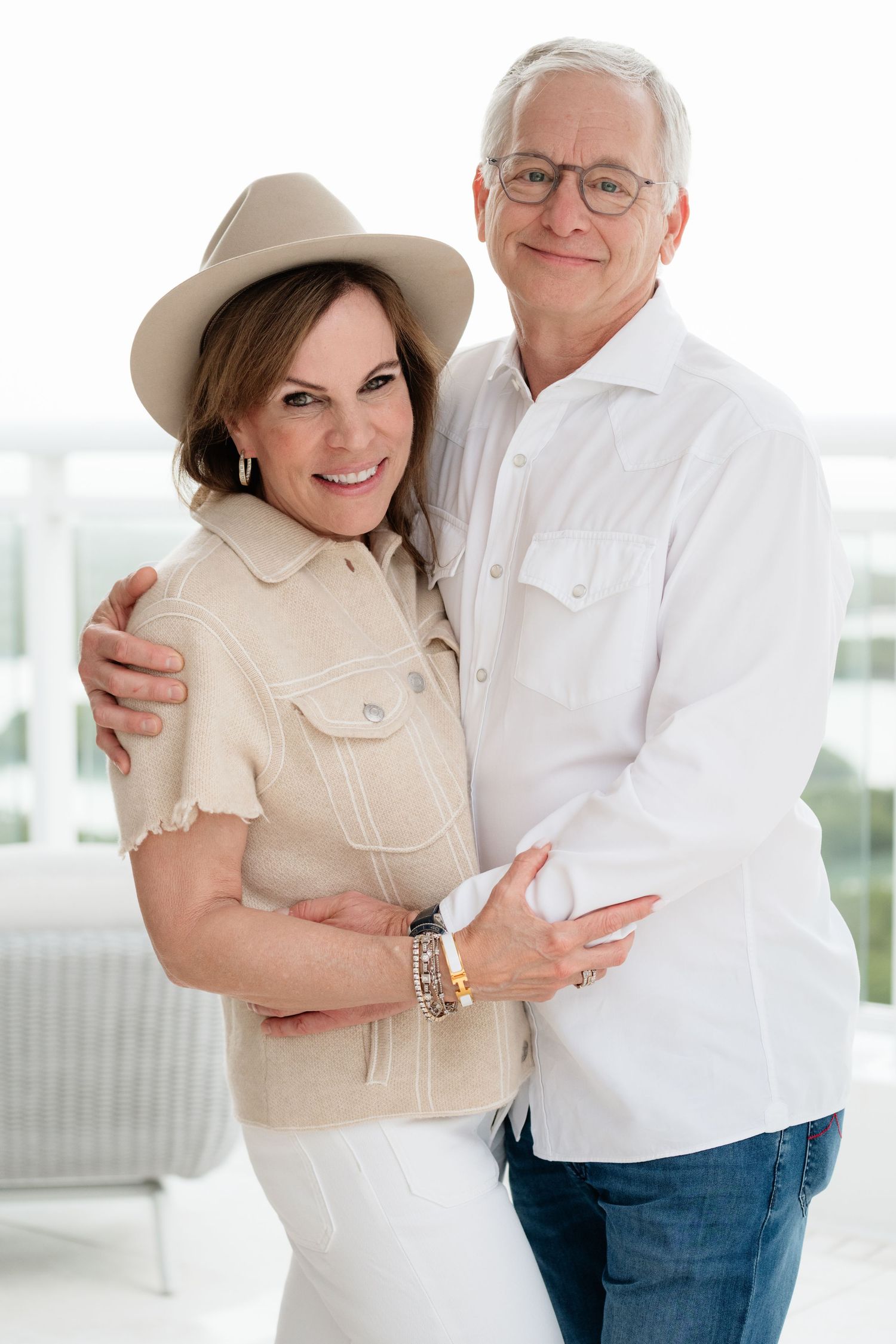 Smiling middle-aged couple embracing outdoors, woman in beige hat and vest, man in white shirt and jeans.
