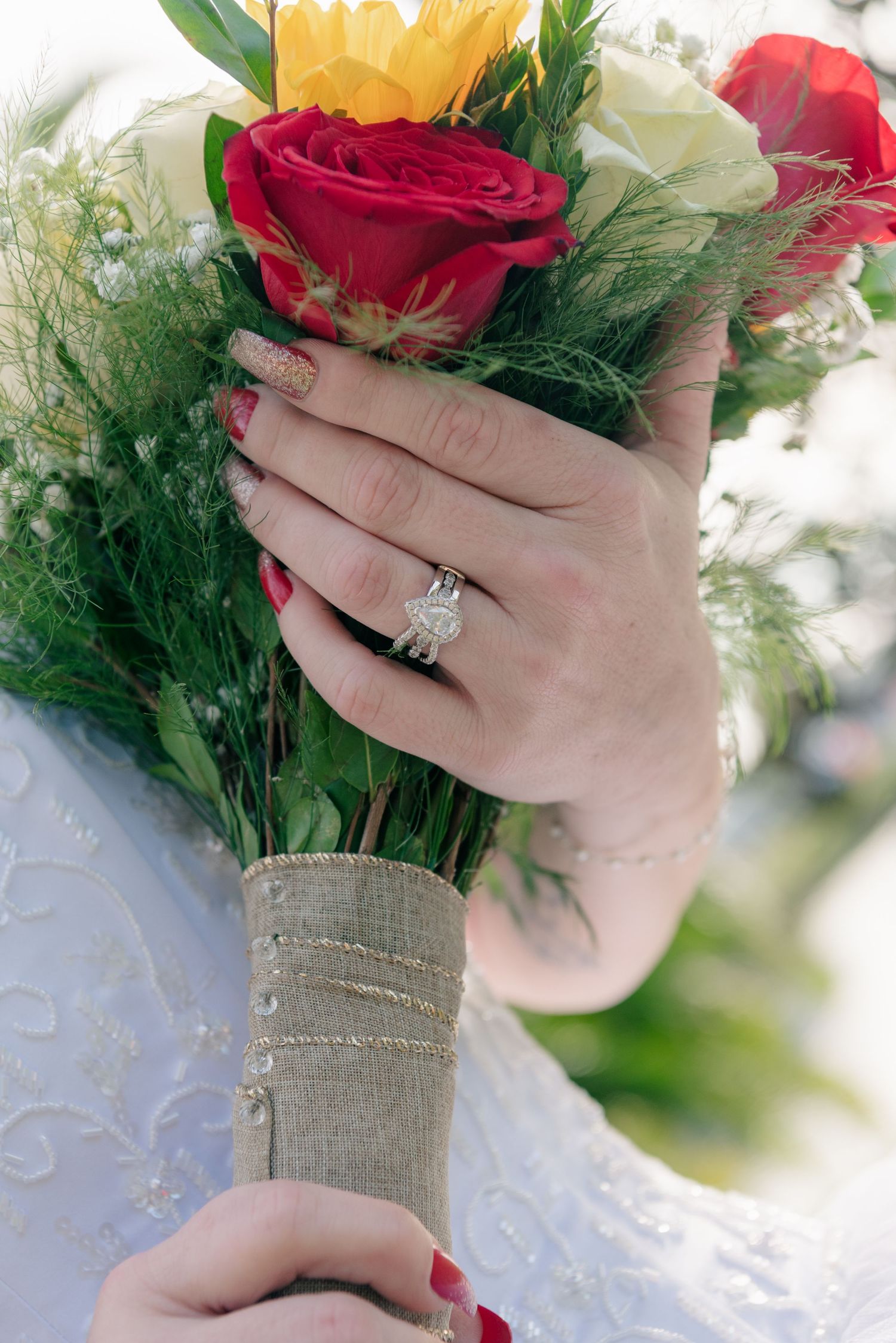 Close-up of hands holding a bridal bouquet with red roses and sunflowers, showcasing an elegant diamond engagement ring.