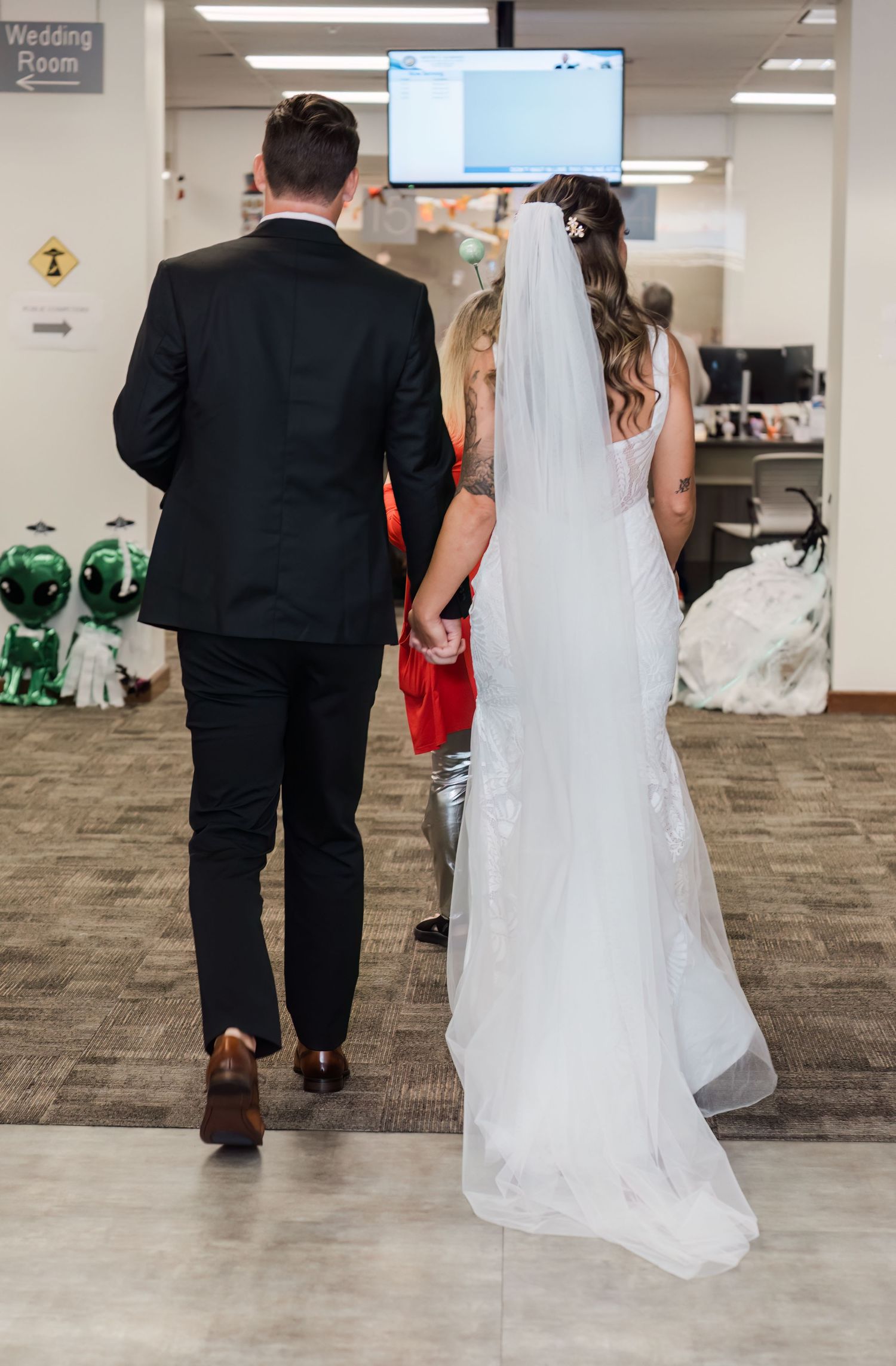 Bride and groom walking hand in hand toward wedding room in an office building, viewed from behind.