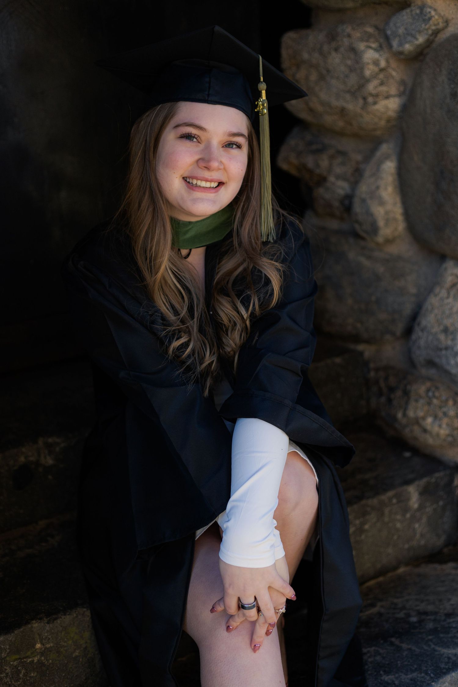 Graduate in academic regalia sits on stone ledge during outdoor photoshoot.