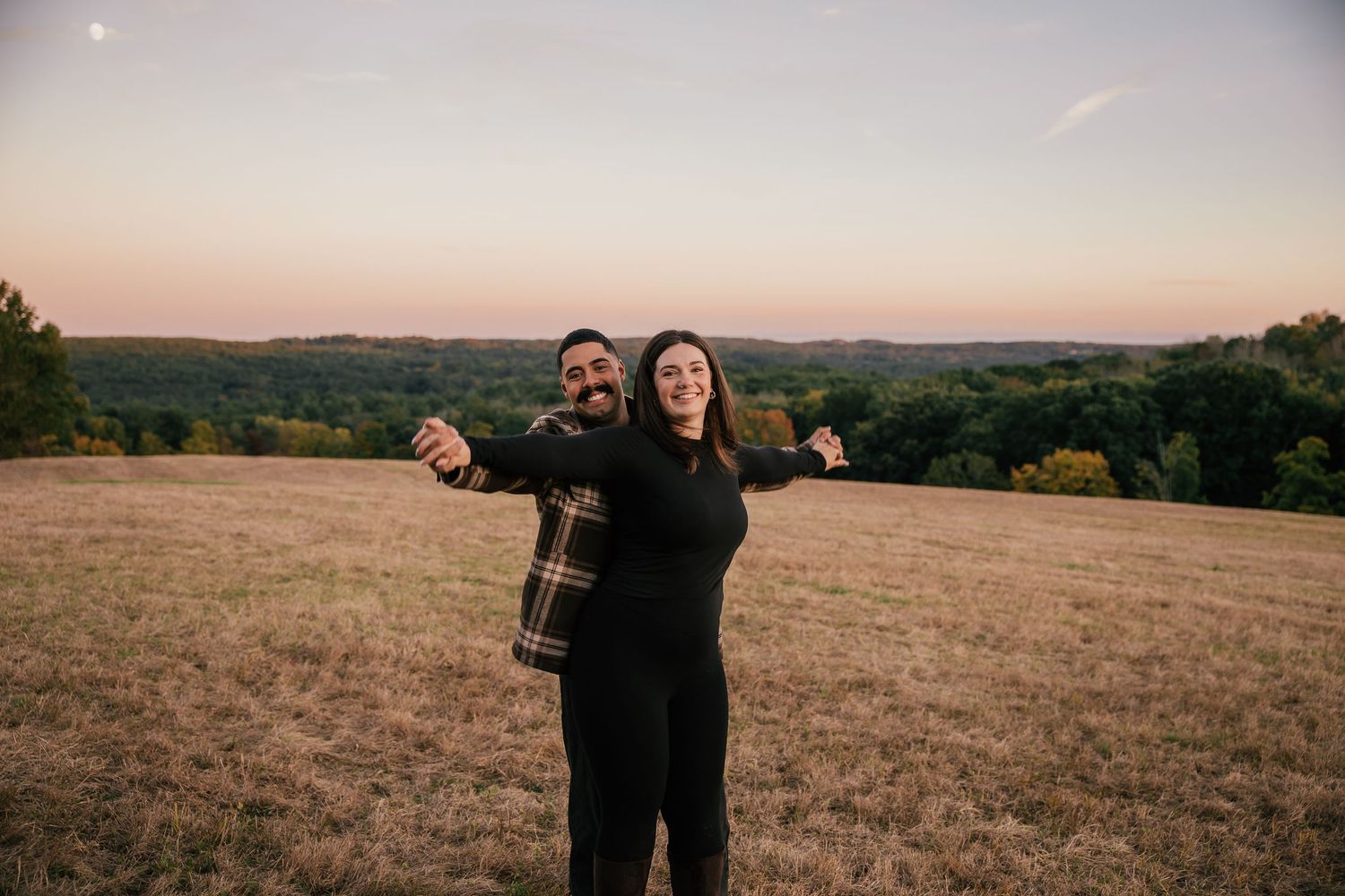 Couple smiling with arms outstretched in golden field at sunset with rolling hills in background.
