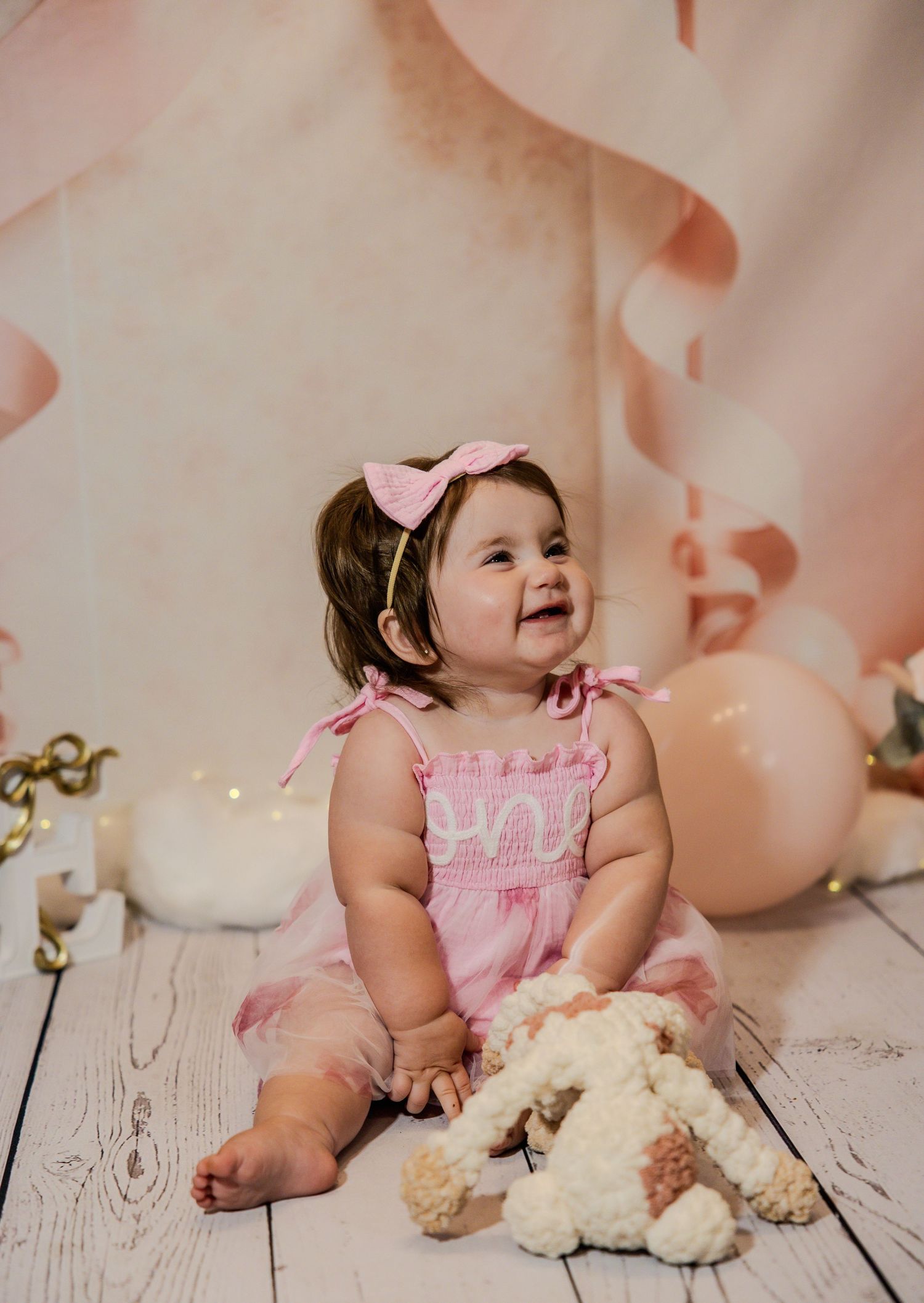 Baby in pink dress sitting on white wooden floor with stuffed animals and pink balloon decorations.