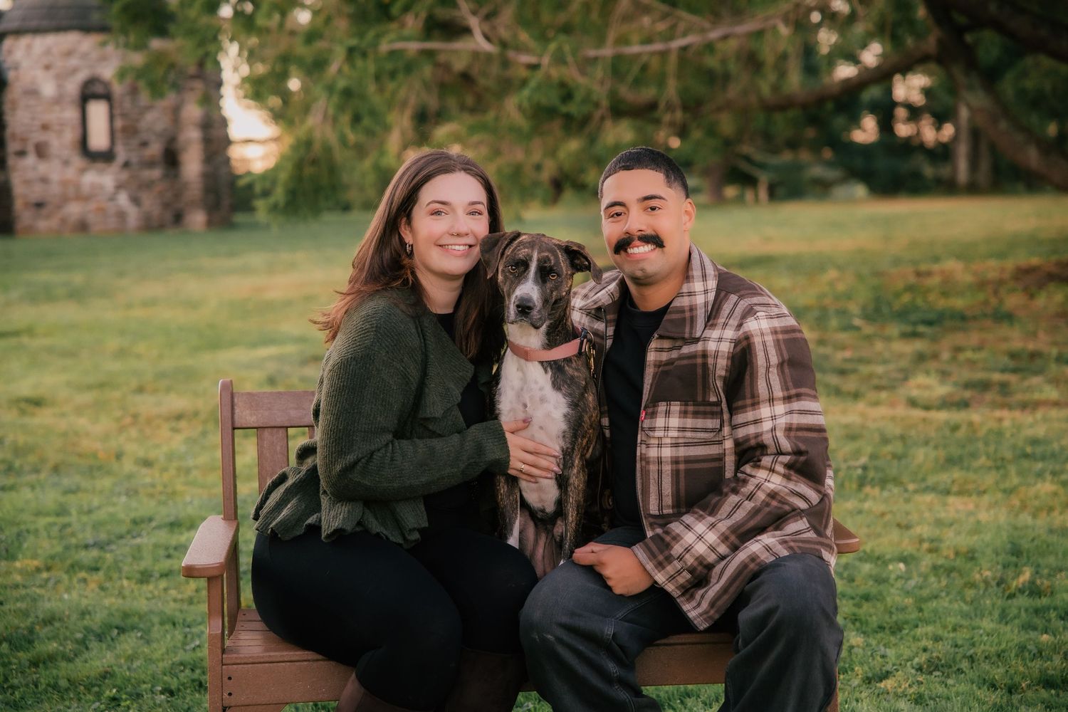 Smiling couple sits on a bench outdoors with their gray dog, surrounded by green grass and trees.
