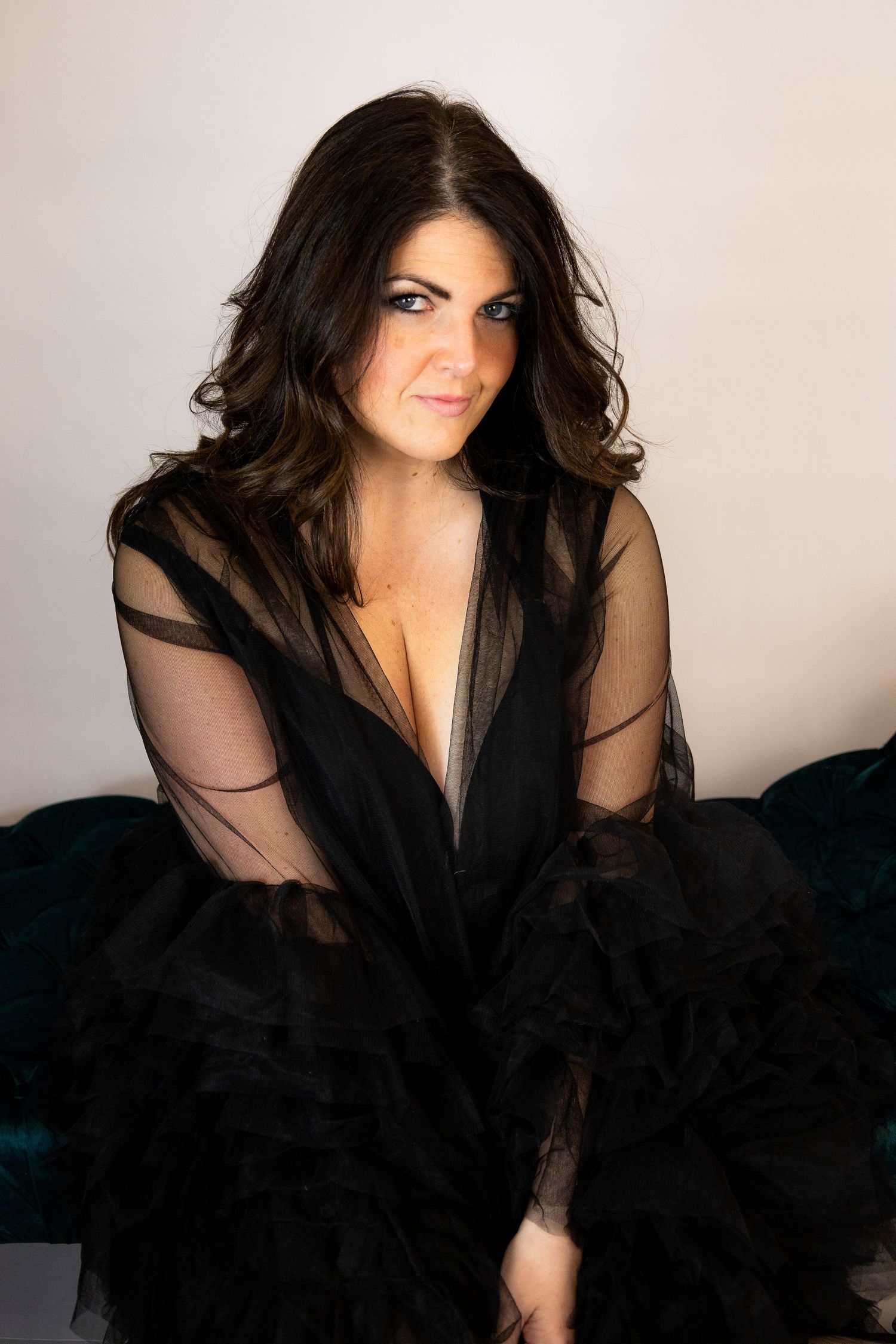 Woman in black sheer tulle robe sitting elegantly on teal velvet chair against white background.