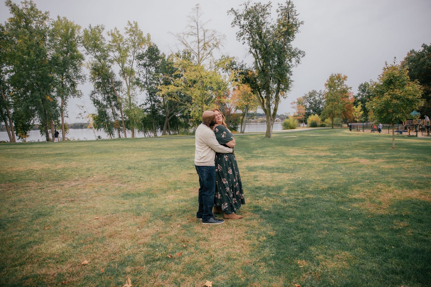 Couple in a loving embrace on a grassy lawn in a park surrounded by tall green trees near a lake during an overcast day.