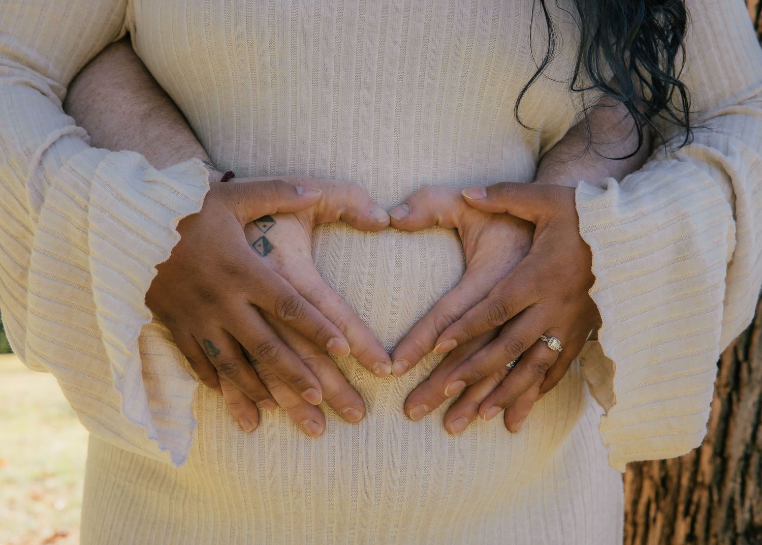 Two hands forming a heart shape on a pregnant woman's belly in a cream ribbed sweater outdoors.