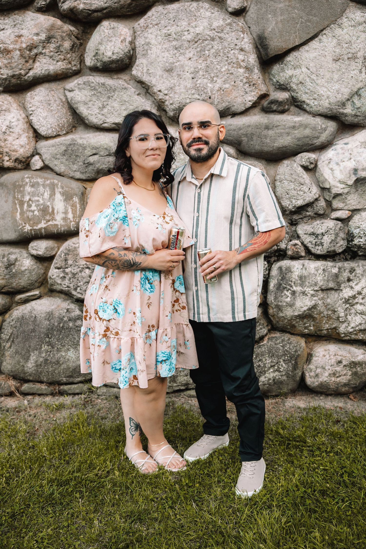 Couple posing together in front of a stone wall, woman in floral dress and man in striped shirt holding drinks.