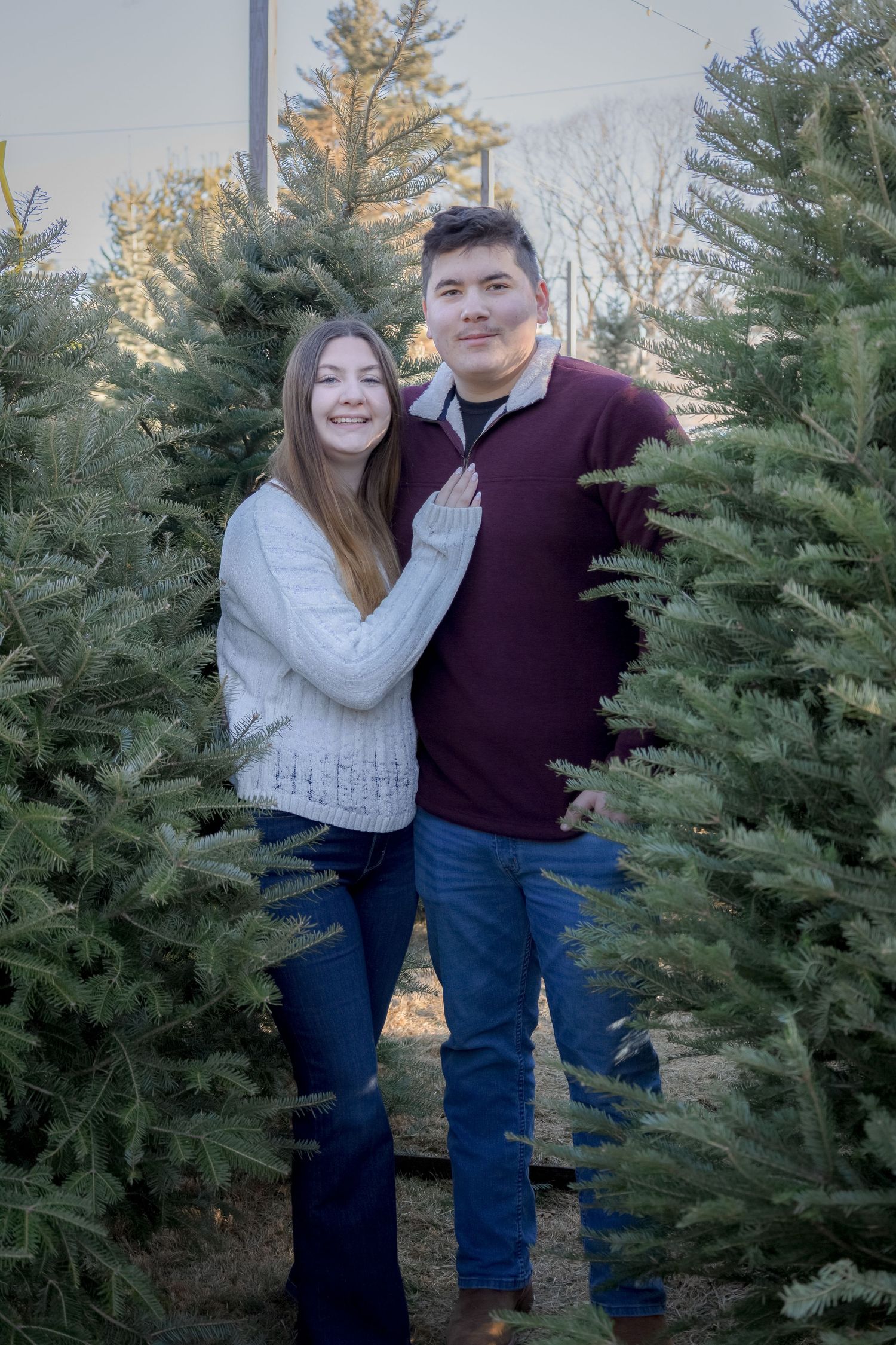 Couple smiling among green Christmas trees at a tree farm, wearing cozy sweaters and jeans in winter.