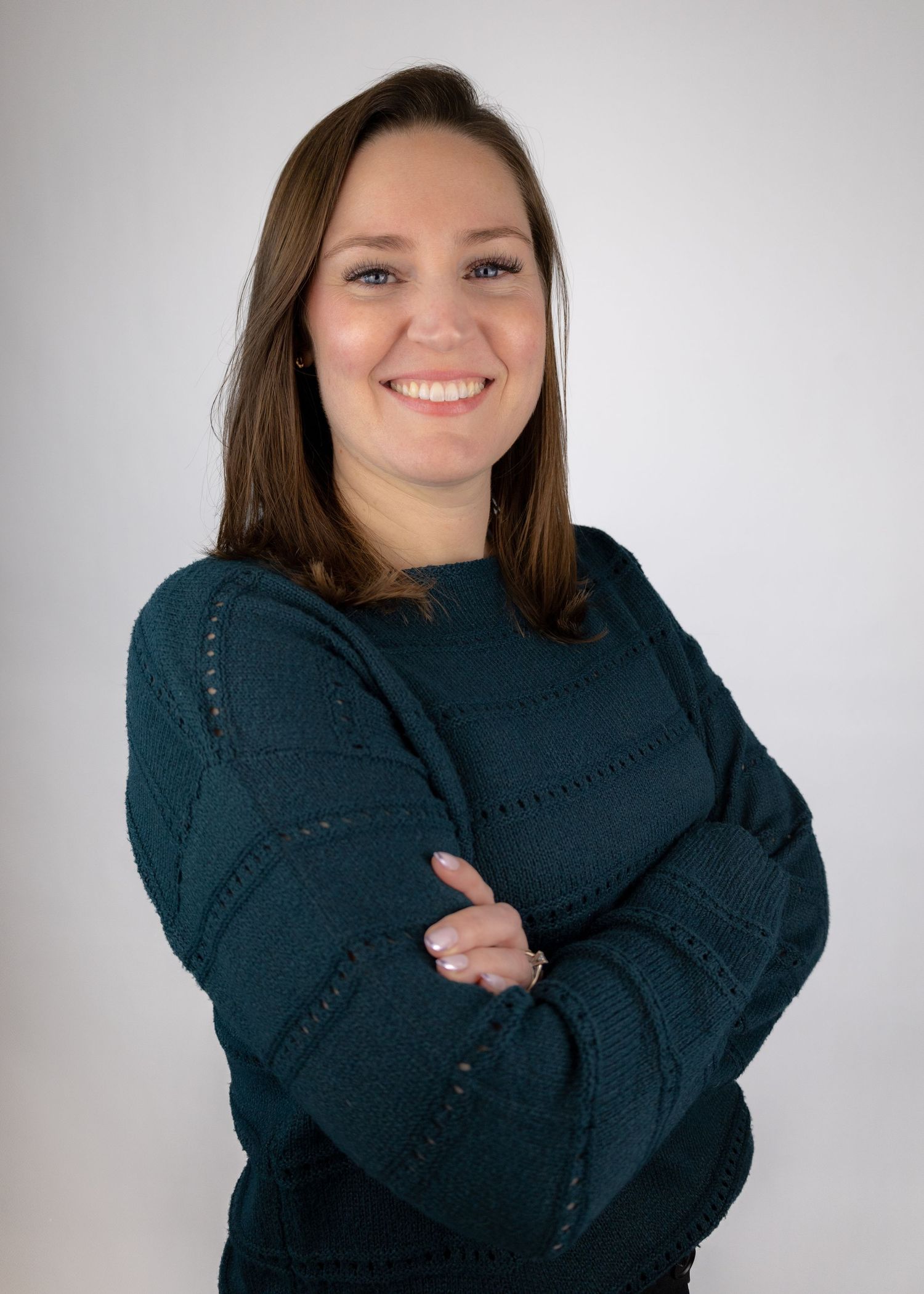 Smiling woman with brown hair in teal sweater posing with arms crossed against gray background.