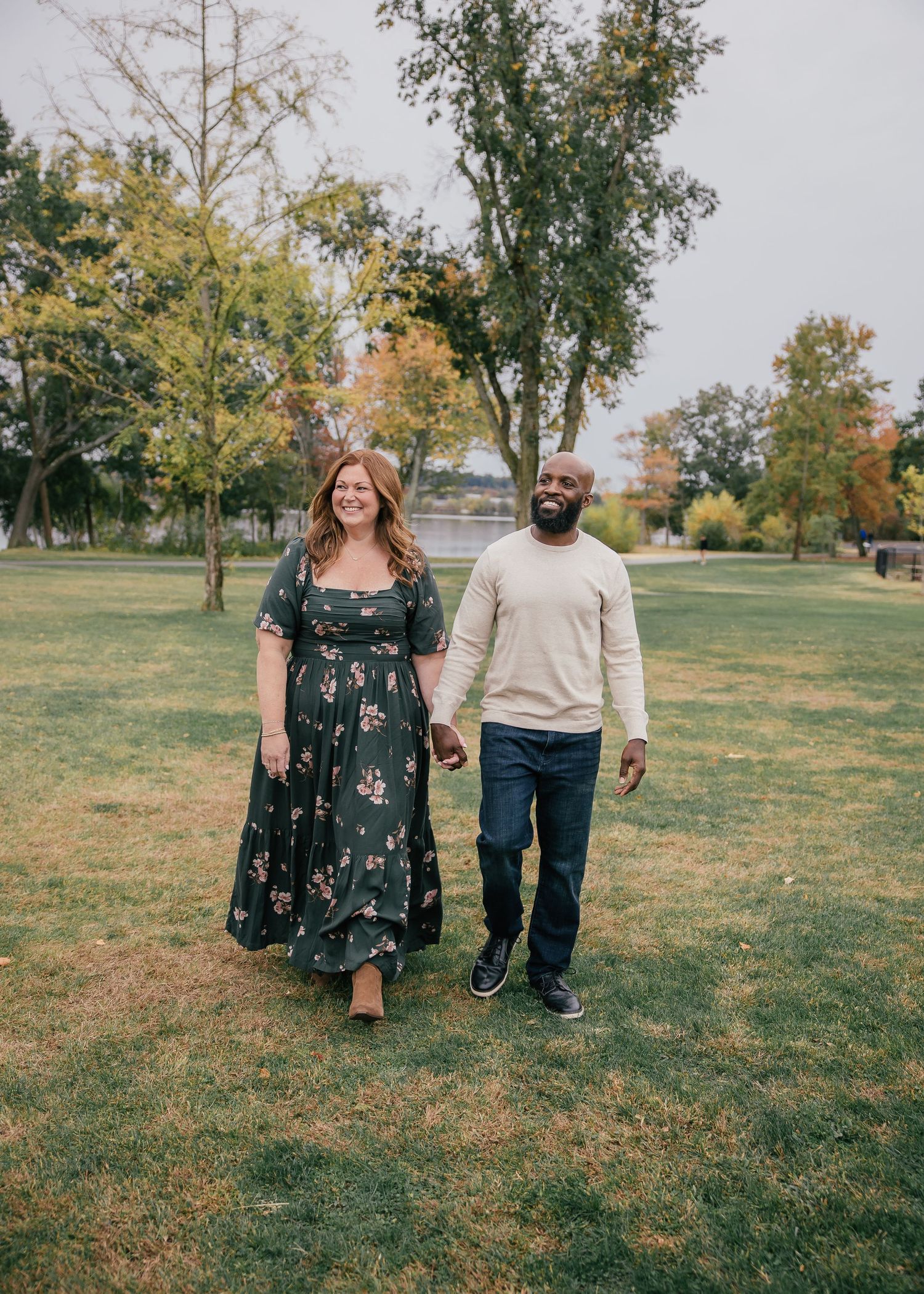 Couple holding hands walking in a green park with autumn trees, woman in floral dress and man in beige sweater.