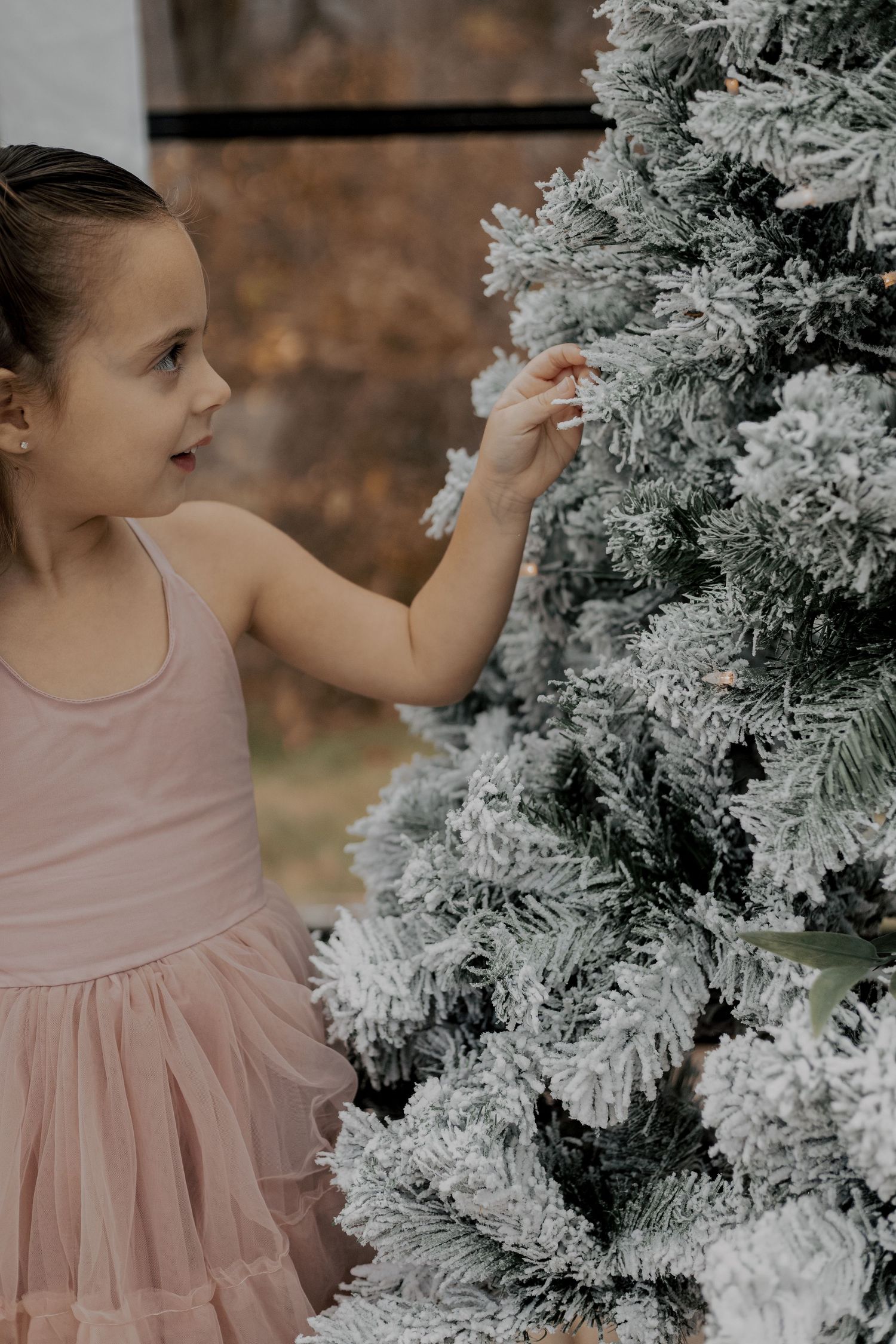 A young girl in a pink tulle dress reaches out to touch a snow-covered Christmas tree.