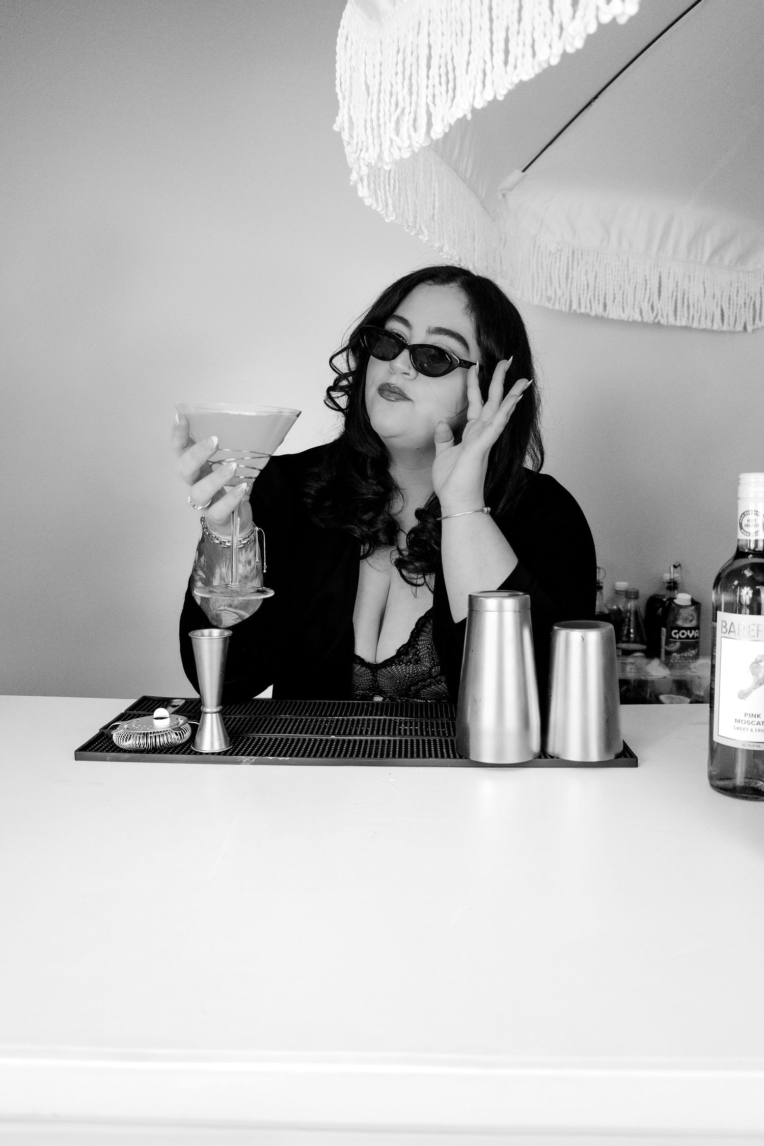 Black and white photo of woman in sunglasses posing at bar with cocktail shakers and bottles.