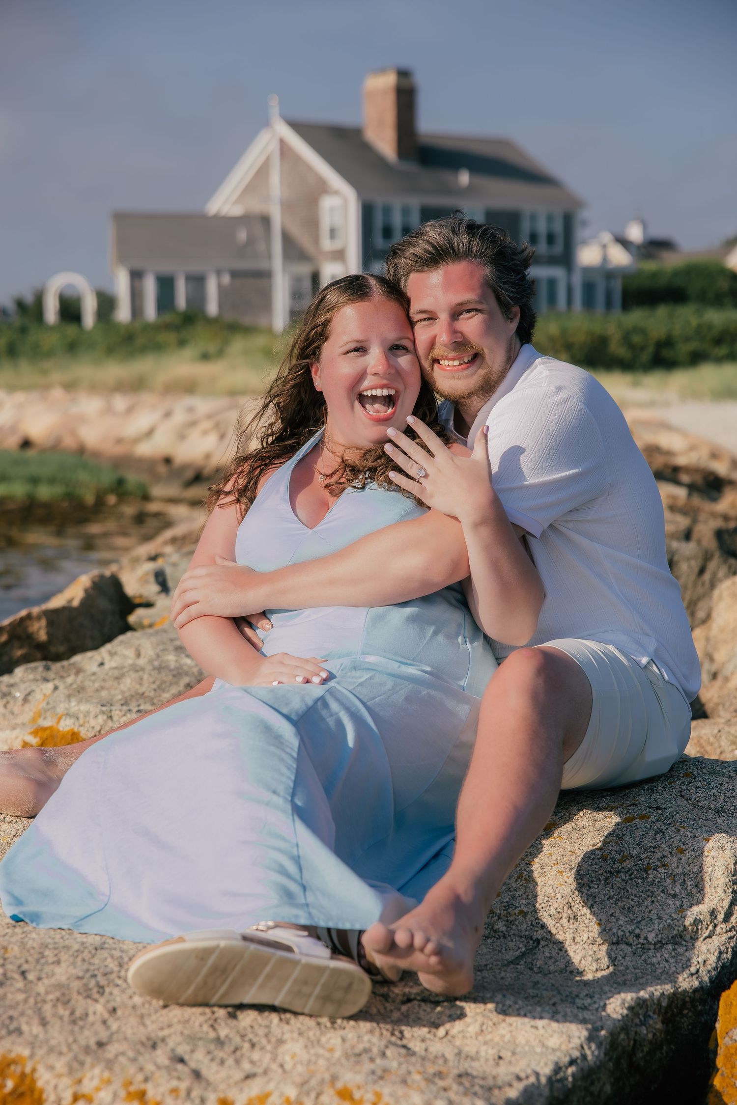 Happy couple sitting on rocky beach, woman showing engagement ring, laughing joyfully together.