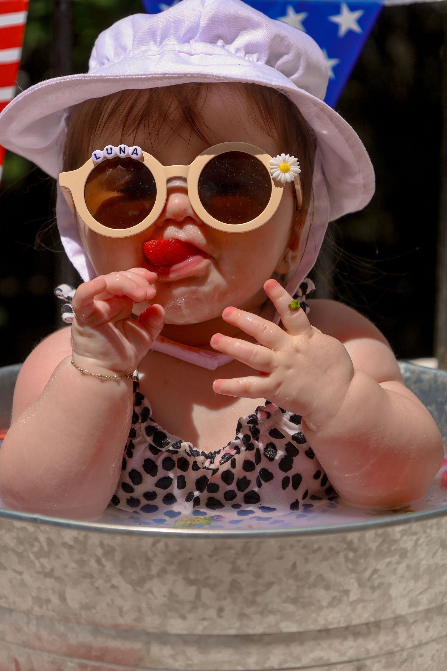 Baby wearing sunglasses and patriotic hat sitting in metal tub during summer celebration.