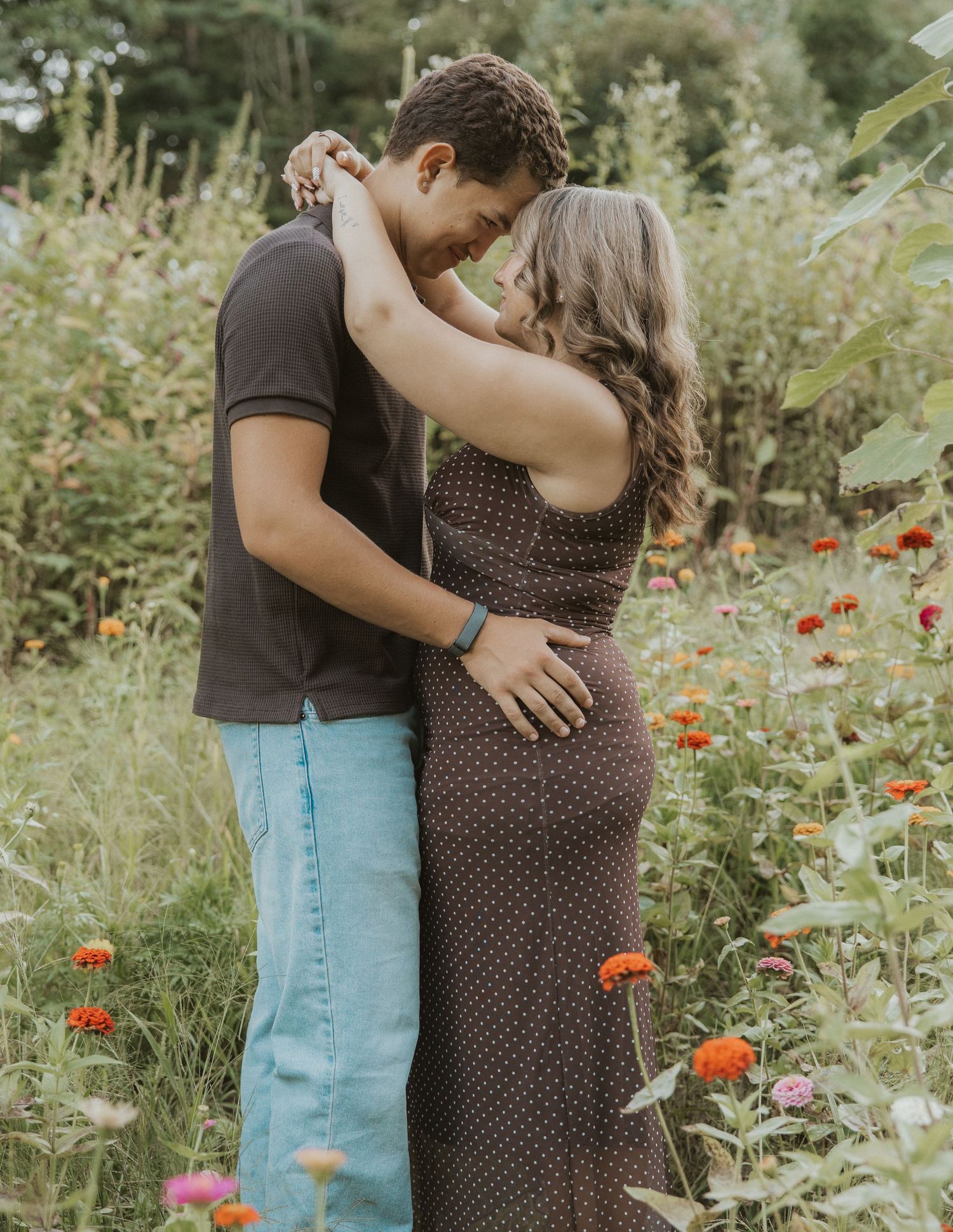 Couple embracing romantically in a colorful flower garden, woman in brown polka dot dress, man in black shirt and jeans.