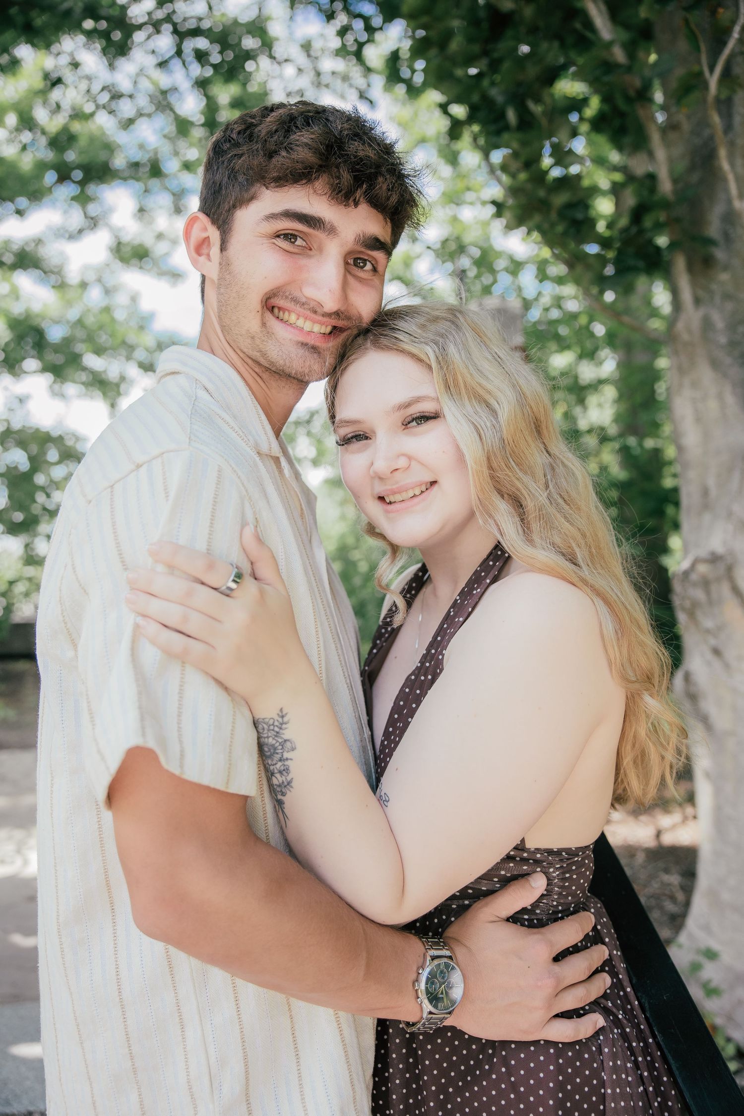 A smiling couple embraces outdoors, woman in polka dot dress and man in striped shirt, with green trees in background.