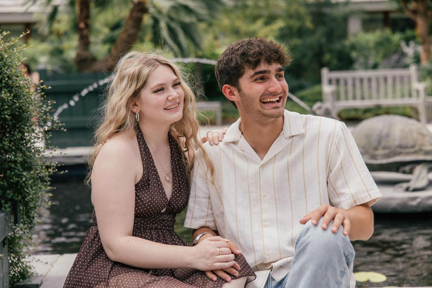 Young couple smiling and sitting together in a lush garden setting near a pond on a sunny day.