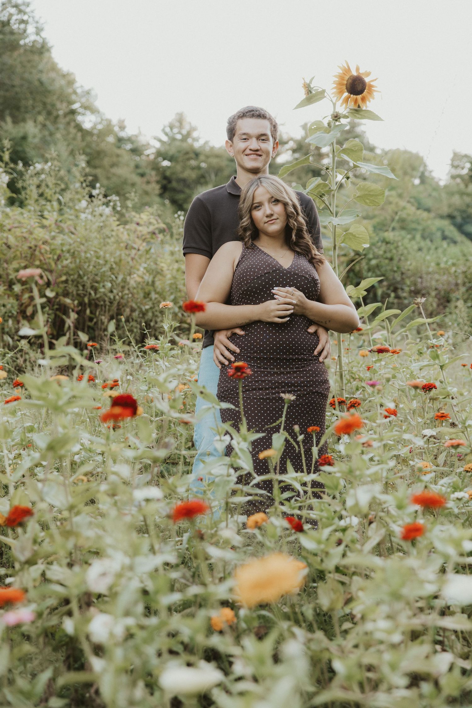 Couple poses in a colorful flower field with sunflowers, man embracing pregnant woman in brown polka dot dress.