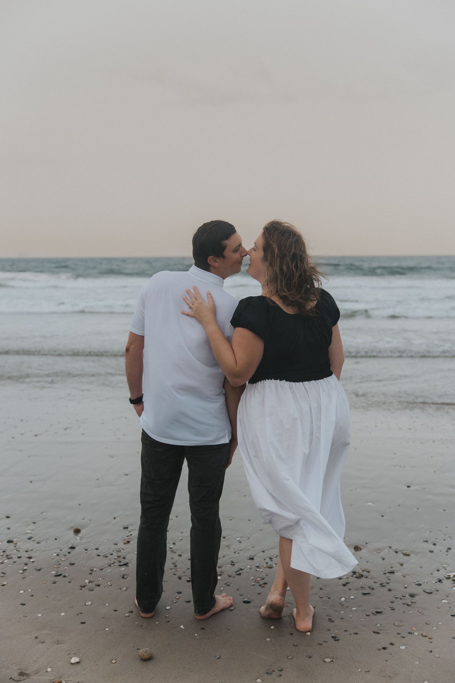 Couple sharing a romantic kiss on a sandy beach at dusk, woman in white skirt and black top, man in white shirt.