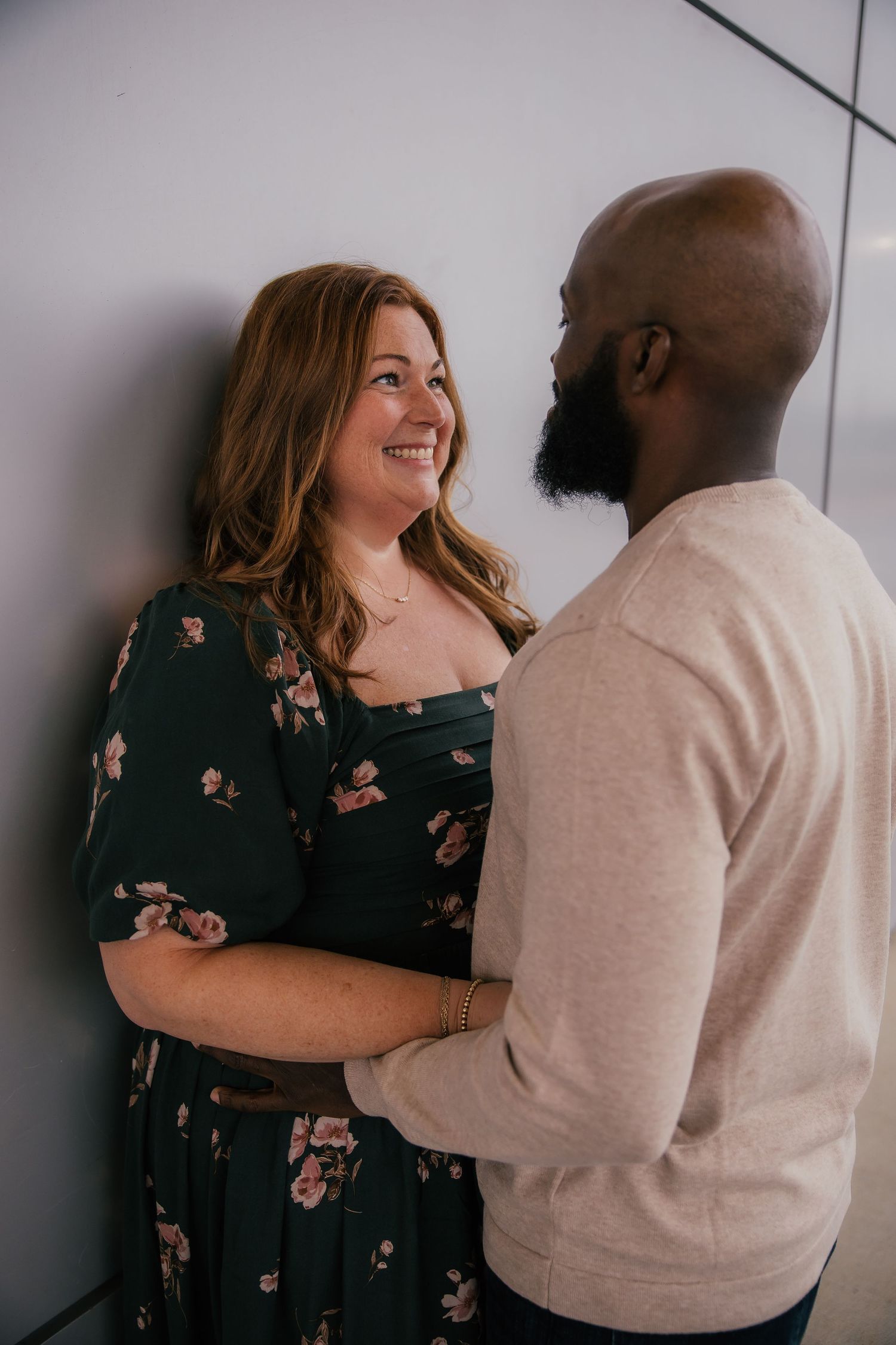 Smiling woman in a dark floral dress embracing a bearded man in a cream sweater during a romantic couple's portrait session.