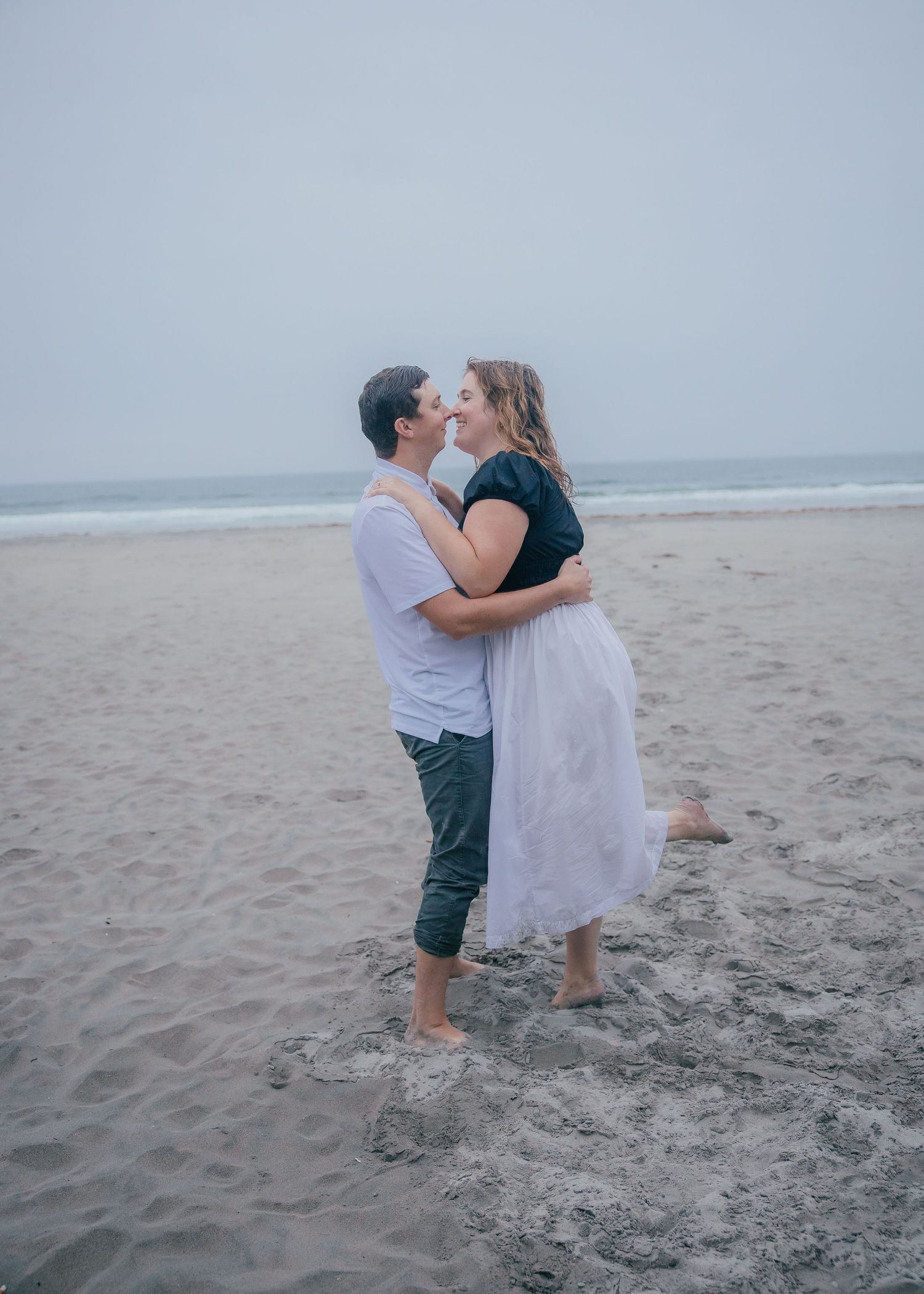 A couple embraces and kisses on a gray, overcast beach, barefoot in the sand with the ocean and waves behind them.