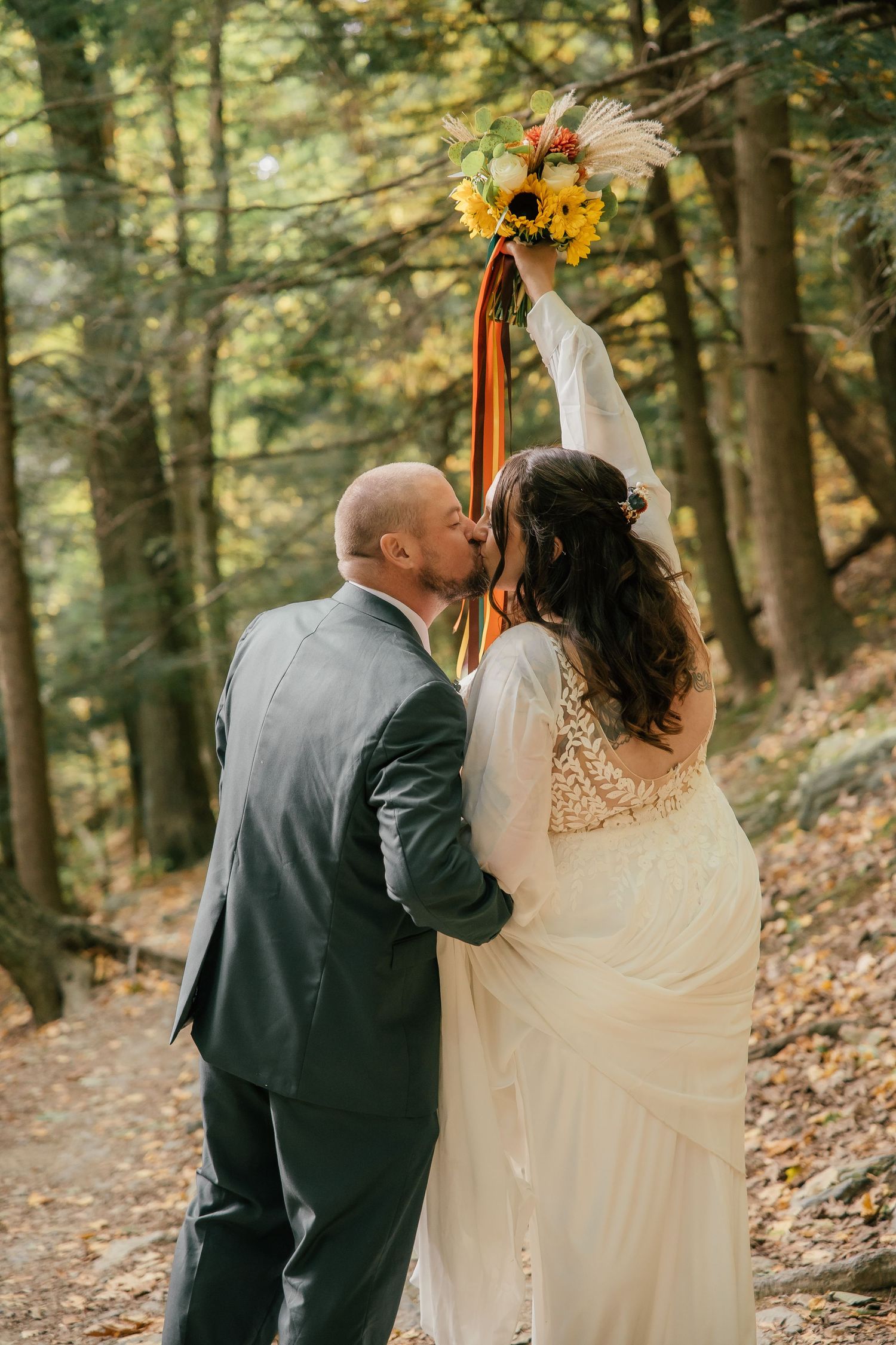 A bride and groom share a kiss in an autumn forest, as the bride raises her sunflower bouquet with colorful ribbons.