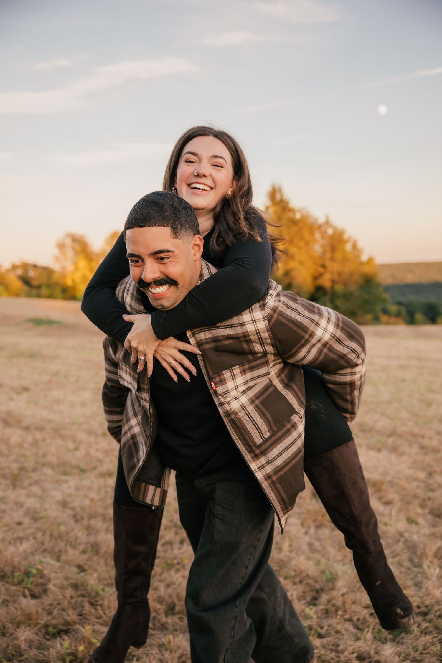 A joyful couple laughing outdoors, man giving woman a piggyback ride in an autumn field at golden hour.
