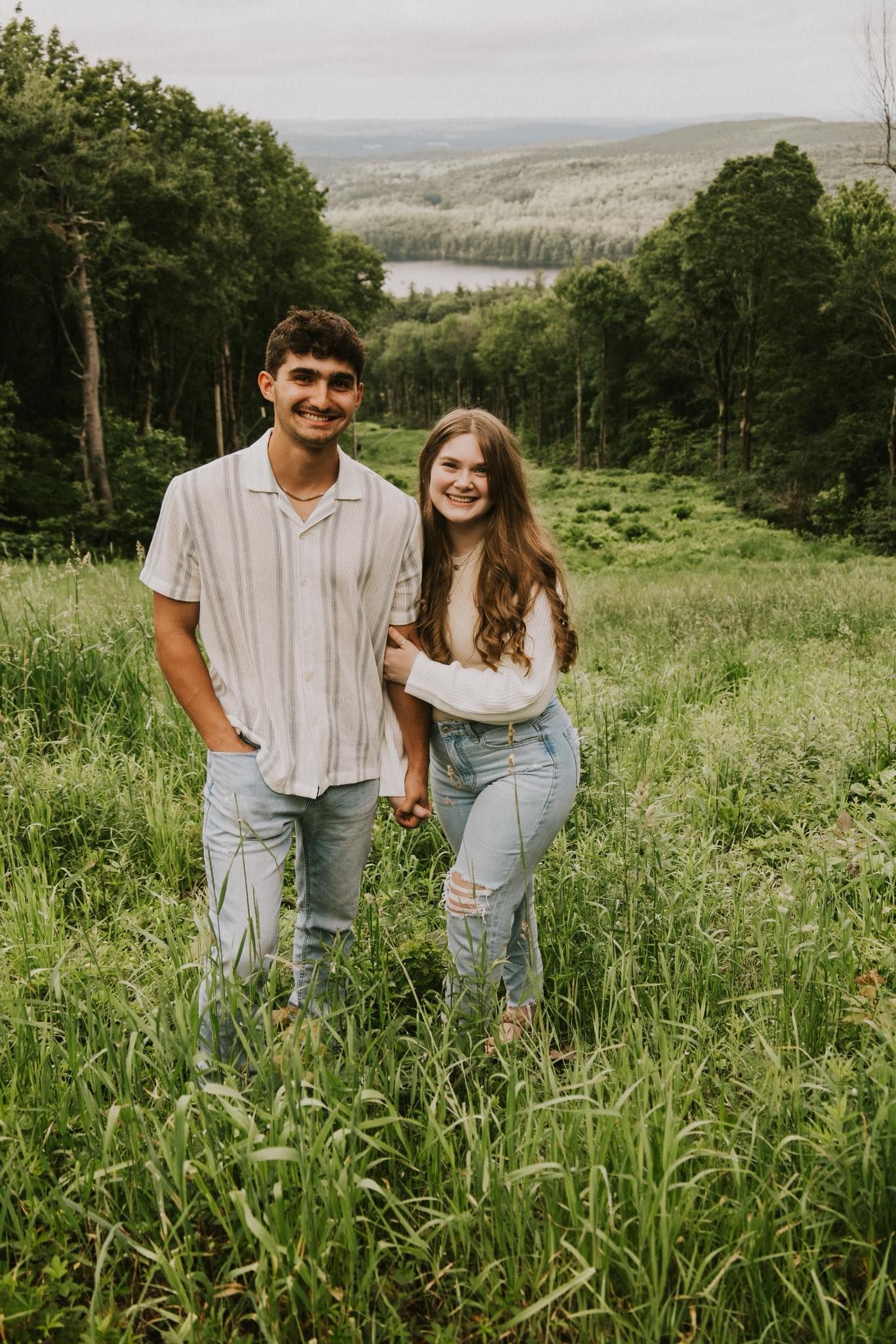 A couple stands together in a lush green meadow with rolling hills in the background during golden hour.