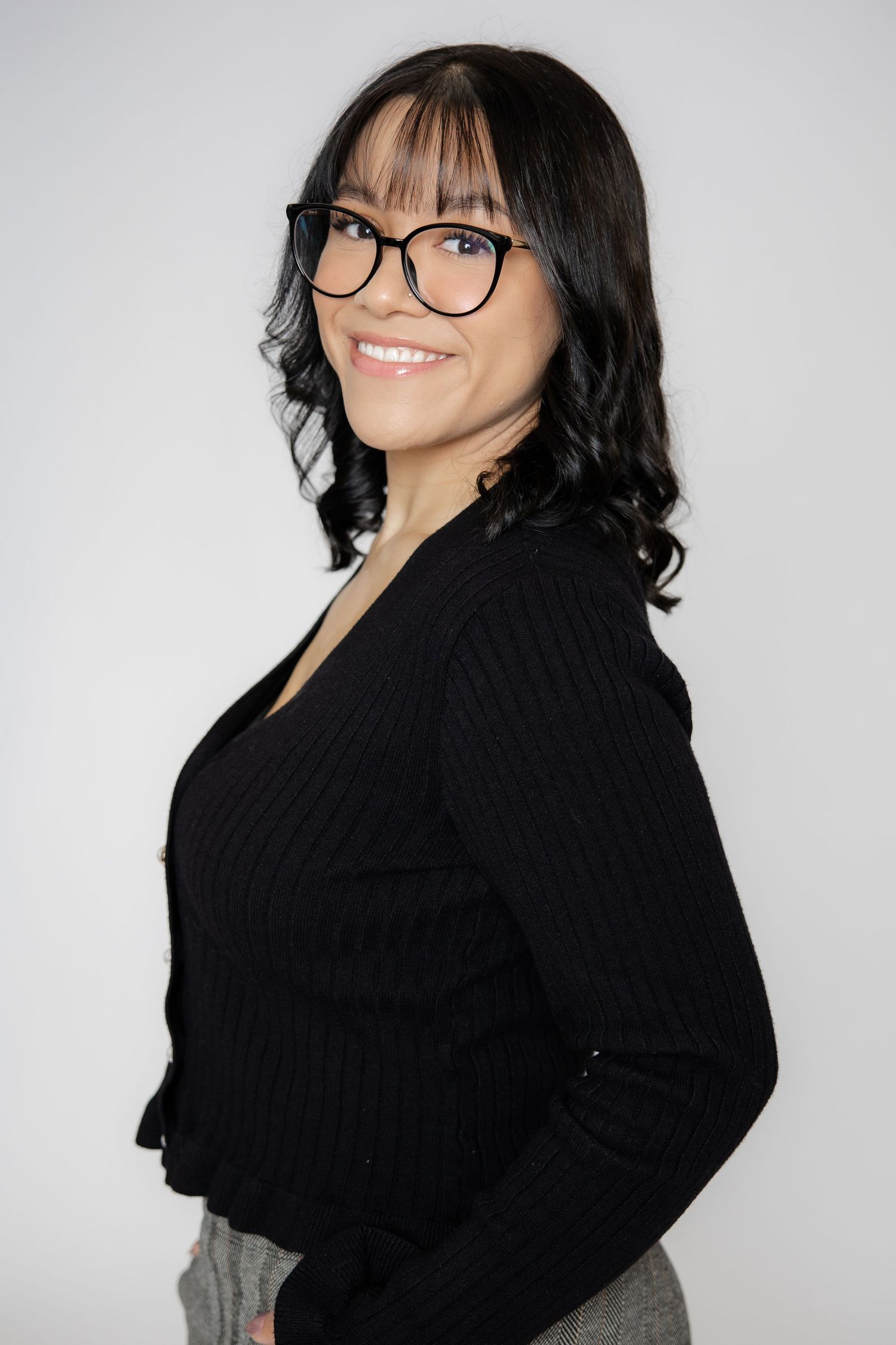 A smiling woman with black glasses and dark wavy hair wearing a black ribbed sweater poses against a white background.