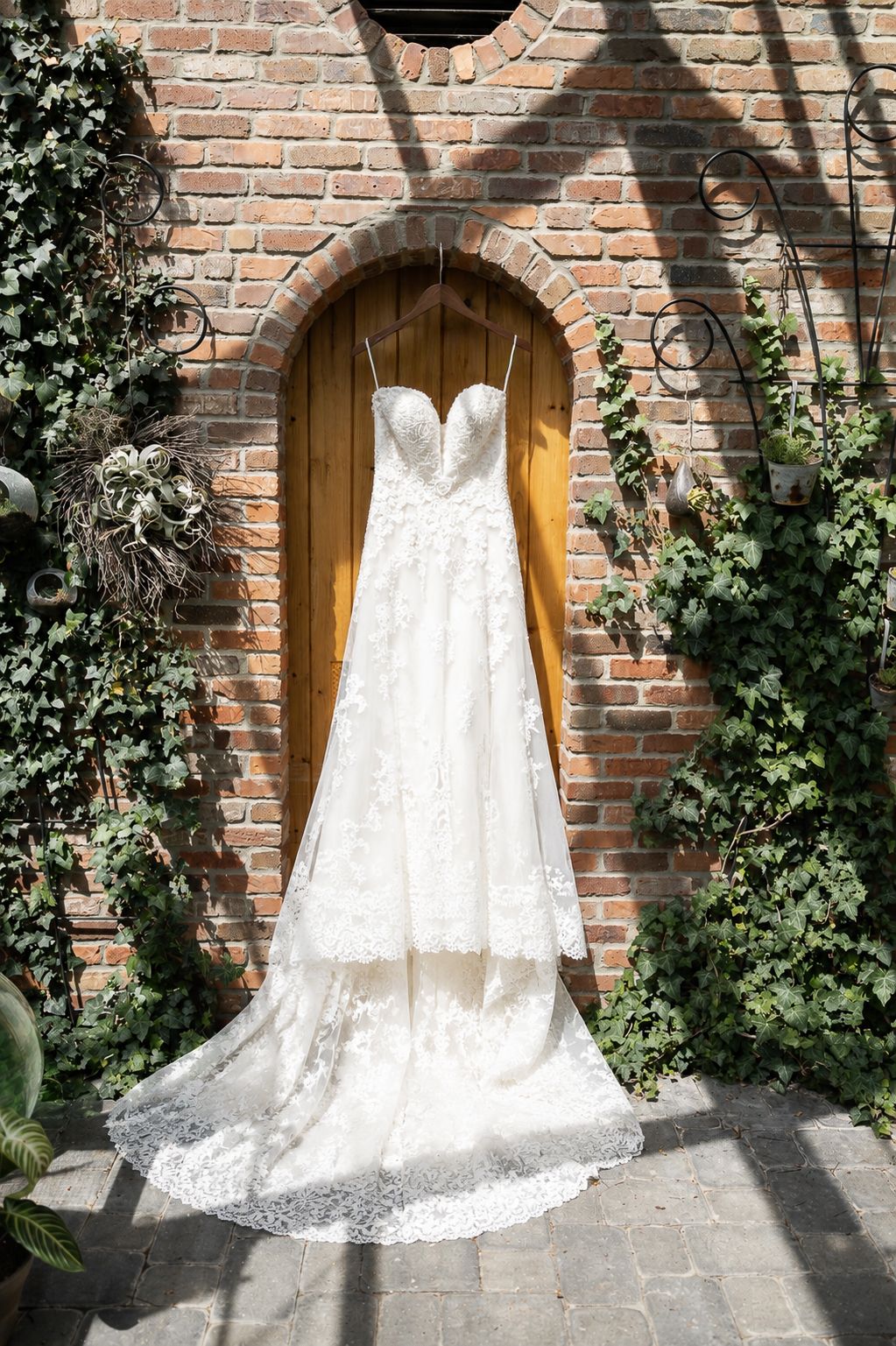 Elegant lace wedding dress hanging on a wooden door framed by brick wall and green ivy.