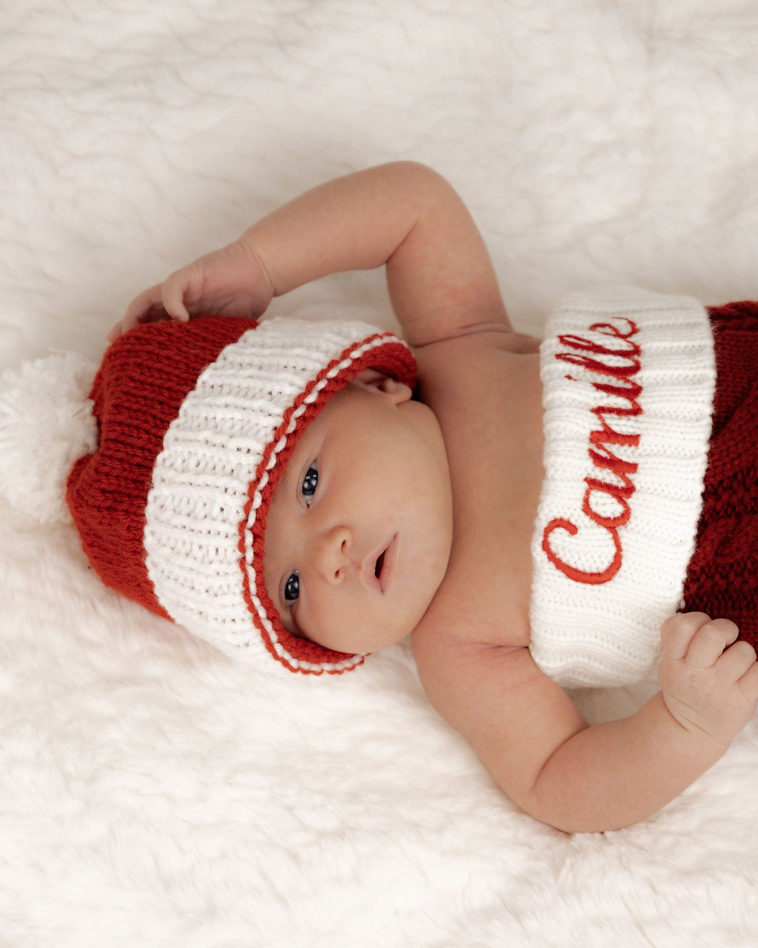 A newborn wearing a red and white knitted Christmas outfit on white fur.